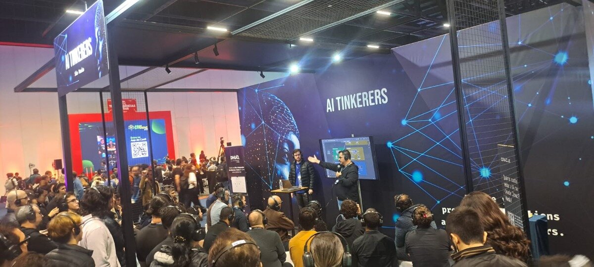 The image captures a technical presentation or workshop session taking place at a large indoor conference or exhibition, featuring a seated audience wearing headphones and two speakers standing in front of a dark blue backdrop labeled 'AI TINKERERS.' The backdrop is decorated with abstract blue network and node graphics.. Text: AI TINKERERS ORACLE DWG Entre na App do [unreadable] pro biens rs. Dr. [unreadable] [other unreadable text]