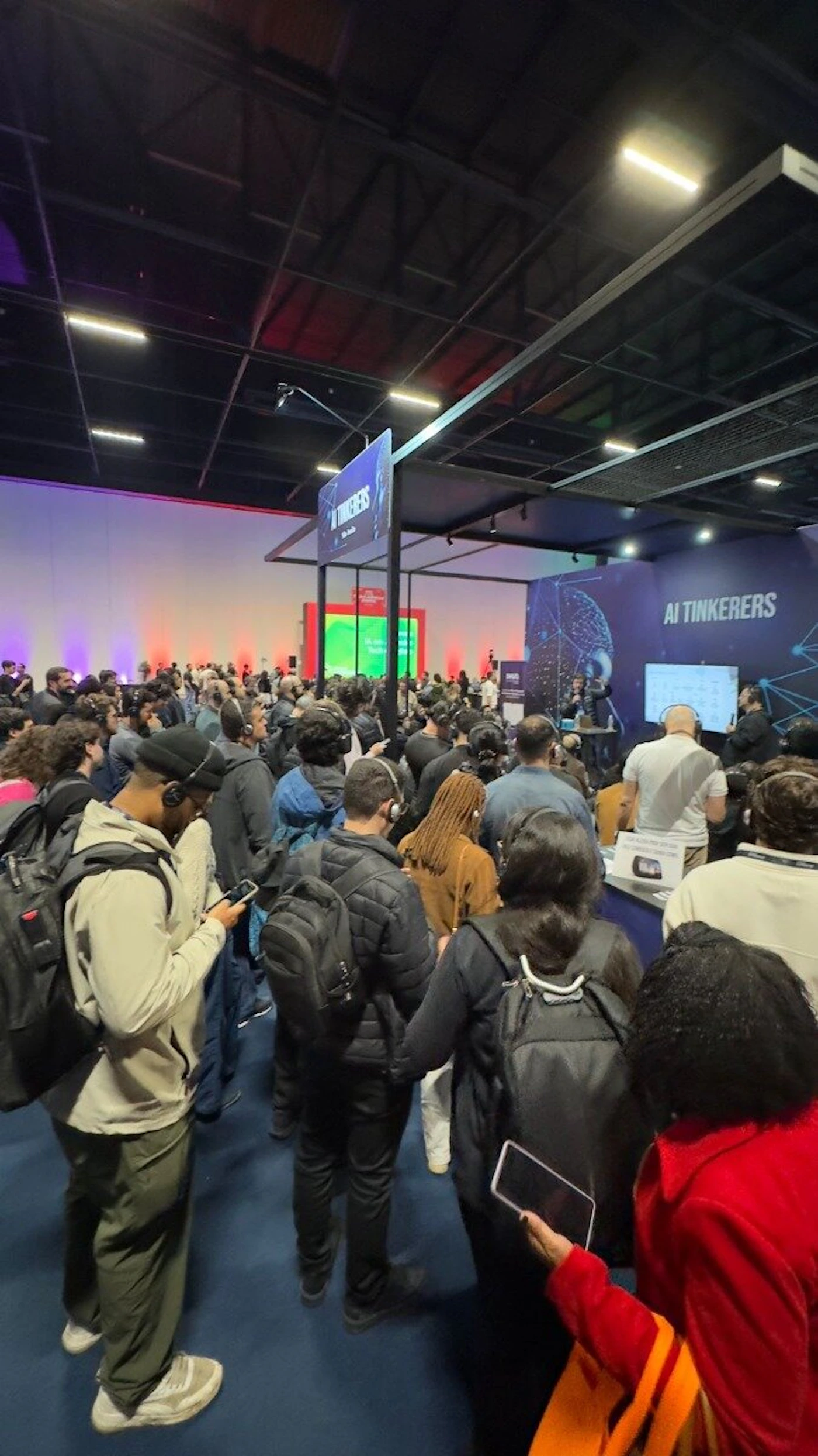 (Photo) A photograph capturing a large crowd of people gathered indoors at a dimly lit conference or event, likely focused on technology or AI. Attendees are facing a presentation area where signage reads "AI TINKERERS." Text: AI TINKERERS A large crowd of people attending a conference or seminar | Indoor, large convention hall or event space | Attendees are wearing headphones; prominent signage for 'AI TINKERERS'; high, dark ceiling structure. | Candid, event photography | Colors: #0A1438, #FFFFFF, #2C2C2C, #800000 Note: The image is a captured moment documenting a real-world event with a large group of people in an indoor setting, fitting the definition of event photography.
