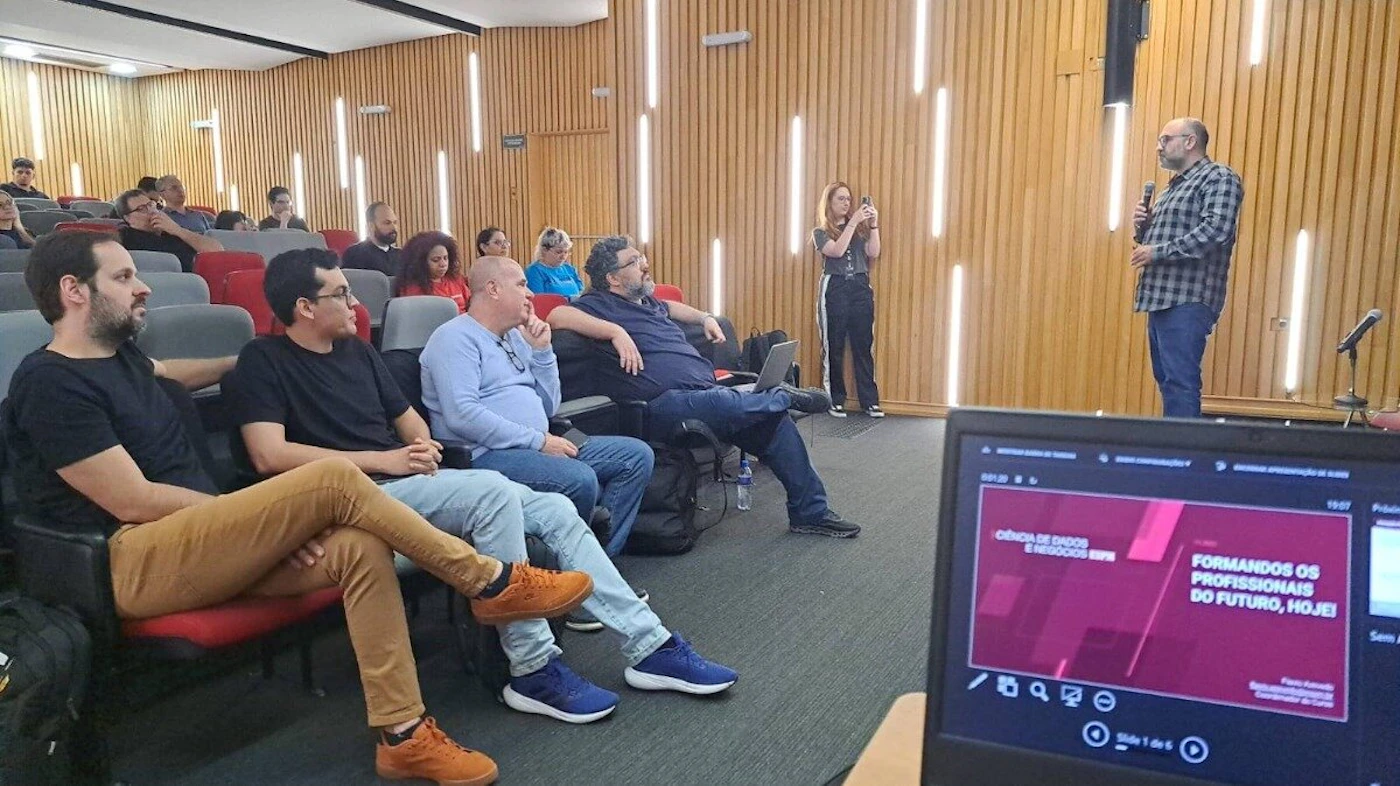 (Photo) This is a photograph of a presentation or lecture taking place in an auditorium with wood paneling and vertical lighting fixtures. Several audience members are visible in the foreground, and a laptop screen displaying the presentation slide is prominent in the bottom right. Text: CIÊNCIA DE DADOS E NEGÓCIOS [unreadable] FORMANDO OS PROFISSIONAIS DO FUTURO, HOJE! [Name] Almeida Coordenador de Cursos Group of people (audience and speaker) attending a professional or academic event. | Indoor, modern lecture hall or auditorium with wooden walls. | Prominent display of a presentation slide on a laptop screen in the foreground; the background features vertical strip lighting embedded in wood-slat walls. | Candid/event photography, taken from the audience perspective. | Colors: #A0522D, #1C1C1C, #8B0000, #F5F5DC Note: The image is a captured photograph of a real-world event—a lecture or presentation—featuring people, an indoor setting, and natural lighting conditions.