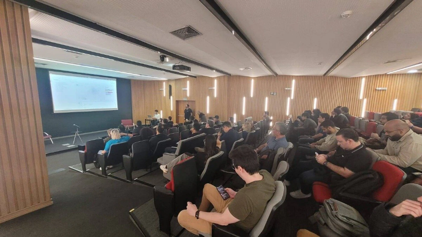 (Photo) A photograph capturing the interior of a modern auditorium or lecture hall during a presentation, showing a large audience seated and facing a projection screen. The room features dark seating, wood-slat wall panels, and vertical strip lighting. Text: [unreadable] A large audience attending a presentation or lecture | Indoor (modern auditorium/lecture hall) | Wood-slat wall paneling, vertical strip lighting, large projection screen, two individuals near the stage. | Candid/documentary | Colors: #404040, #B8A888, #FFFFFF Note: The image is a captured moment of a real-life event, specifically showing people gathered in an indoor setting for a lecture or conference, which classifies it as a photograph.
