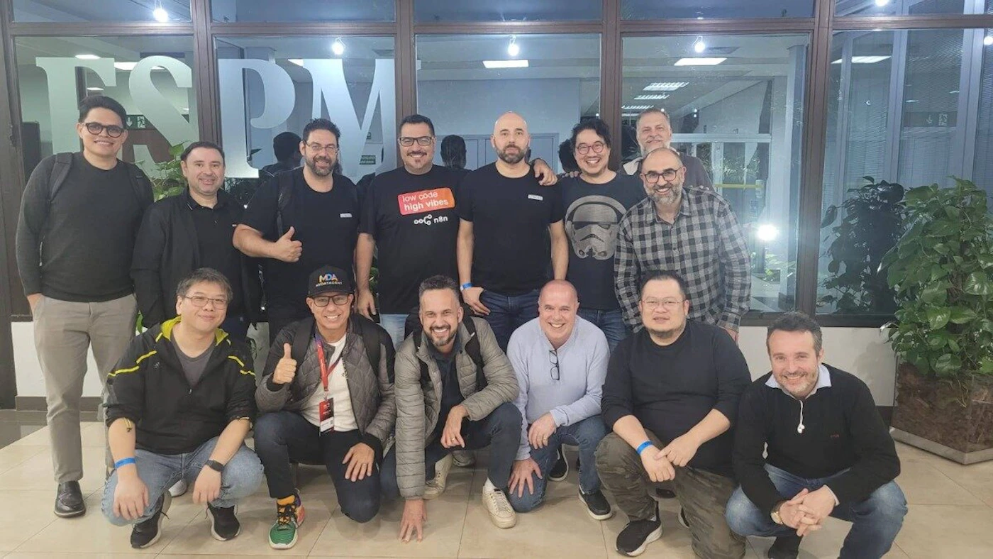 (Photo) A group photograph features approximately fourteen men posing together indoors, standing and crouching in front of large glass windows that display the letters FSPM. Text: FSPM low code high vibes MQA Group of men (colleagues or event attendees) | Indoor, likely a lobby or hallway, with large windows visible in the background. | The group is composed entirely of men; large letters (FSPM) are visible on the glass windows behind them; one man is wearing a t-shirt with a Star Wars Stormtrooper design. | Group portrait/candid | Colors: #000000, #808080, #D3D3D3, #4F4F4F Note: This is a standard photographic capture of a group of people in a real-world, indoor setting, characteristic of a group portrait.
