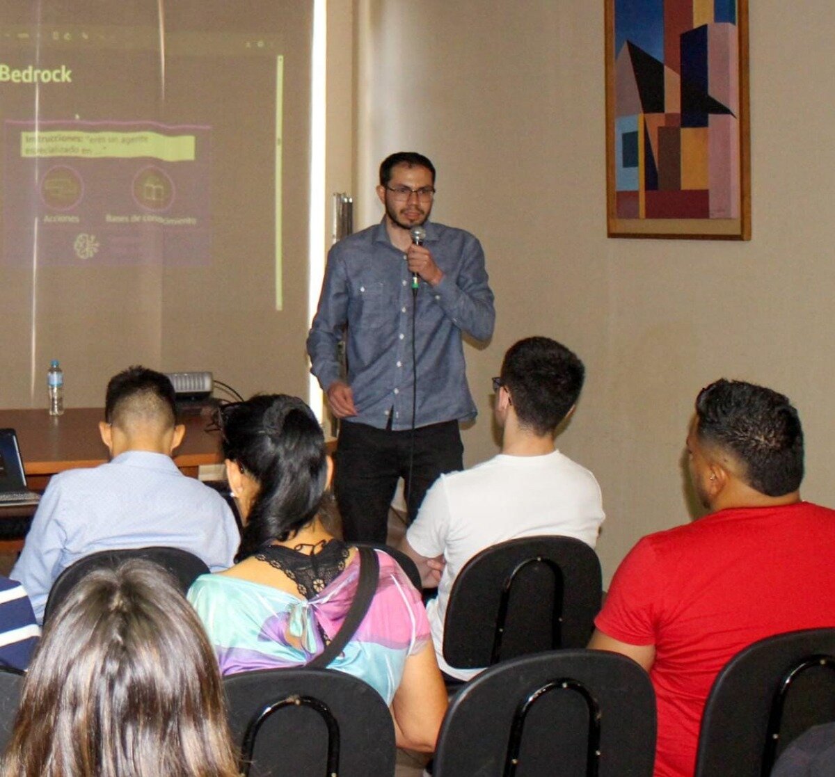 (Photo) A photograph capturing a speaker addressing an audience in an indoor setting, with a presentation slide projected onto a screen behind him and an abstract geometric painting hanging on the wall. Text: Bedrock las transacciones: ¿eres un agente es especializado en...? Acciones Bases de conocimiento People (speaker and audience) | indoor (conference room or classroom) | Speaker holding a microphone, projected presentation slide visible, abstract artwork on the wall. | Candid/Event documentation | Colors: #D9D1C7, #4A6E94, #E82929, #4A004A Note: The image is a realistic capture of a real-world event, documenting a speaker and an audience during a presentation.