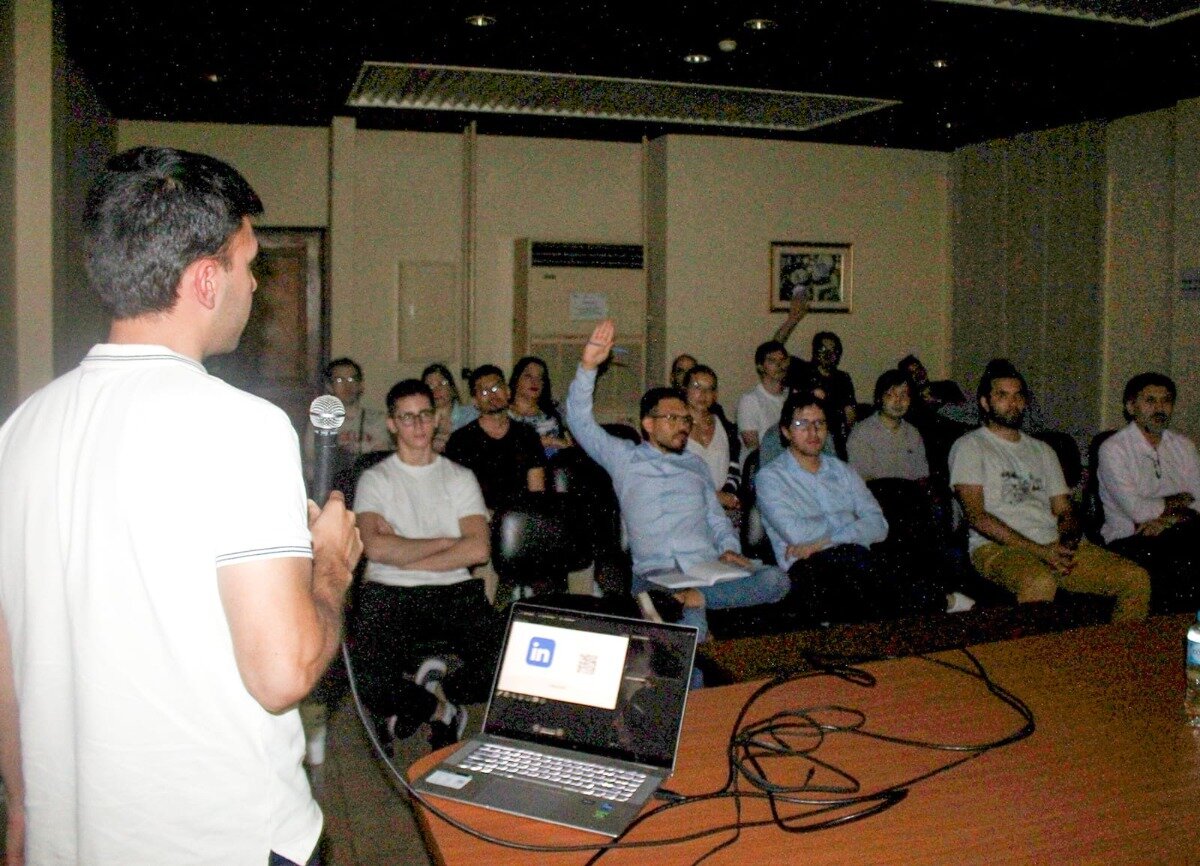 (Photo) A photograph capturing a presentation or meeting in a dimly lit room, showing a speaker addressing an audience, one member of which has raised his hand to ask a question. Text: in A speaker and an audience during a professional or educational presentation. | Indoor, conference room or lecture hall. | The speaker holds a microphone; a laptop on the table displays the LinkedIn logo; an audience member is raising his hand. | Candid, documentary style, low light. | Colors: #000000, #FFFFFF, #A0522D, #336699 Note: This image is a capture of a real-life event featuring people in an indoor setting, characteristic of a photograph.