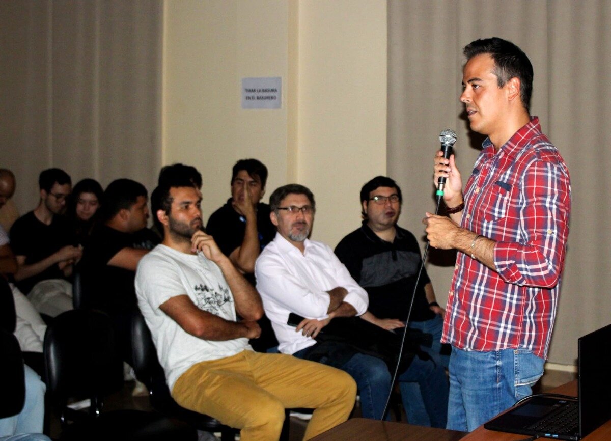 (Photo) A man in a red and white plaid shirt is standing, holding a microphone, and speaking to a seated audience in an indoor setting, likely during a presentation or lecture. Text: TIRAR LA BASURA EN EL BASURERO Group of people (speaker and audience) | Indoor (conference room or classroom) | Speaker holding a microphone; audience members seated and listening; a small instructional sign is visible on the wall. | Candid/event photography | Colors: #A82424, #E6DED0, #404040, #C8A040 Note: The image is a realistic photograph documenting an event, showing people in an indoor setting, which fits the definition of a 'photo'.