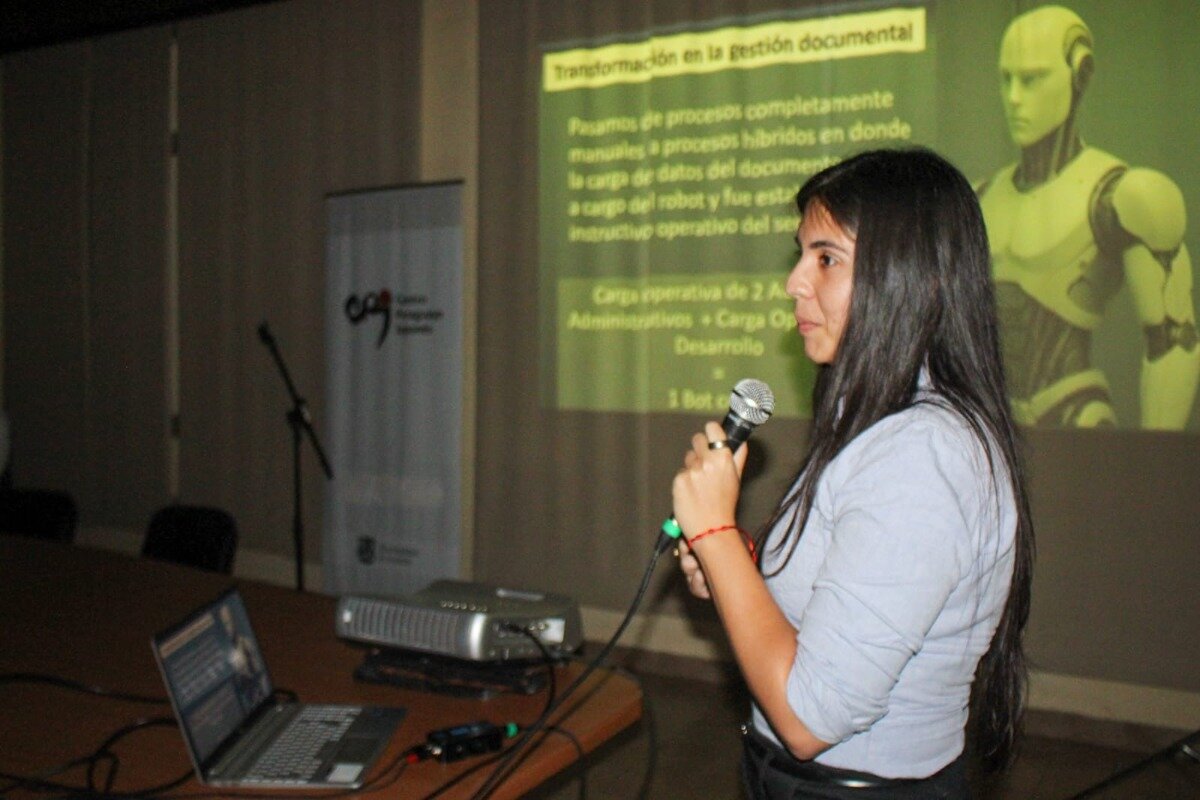 (Photo) A woman is giving a presentation in a dimly lit conference room, speaking into a microphone while standing in front of a projected screen that displays Spanish text about document management and a 3D rendering of a humanoid robot. Text: Transformación en la gestión documental Pasamos de procesos completamente [unreadable] manuales a procesos híbridos en donde la carga de datos del document[o] a cargo del robot y fue esta instructivo operativo del se[rvicio] Carga operativa de 2 A[dministrativos] + Carga Op[erativa] Desarrollo 1 Bot [unreadable] Person (presenter) and technology (presentation slide, projector, laptop) | Indoor (conference room/lecture hall) | Speaker holding a microphone; large projected slide with AI/robotics theme; dim lighting; visible banner/signage on the left. | Candid/event photography | Colors: #333333, #A9BCCF, #8BC34A, #FDD835 Note: The image is a photograph capturing a real-world event, specifically a speaker giving a presentation in a conference or lecture setting.