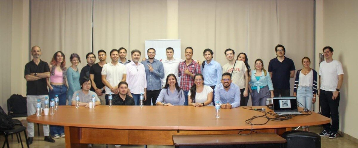 A group photo captures approximately two dozen individuals, both men and women, posing behind a long, curved wooden conference table in an indoor setting. The background consists of light-colored, floor-to-ceiling curtains, and a laptop displaying a QR code is visible on the right side of the table.