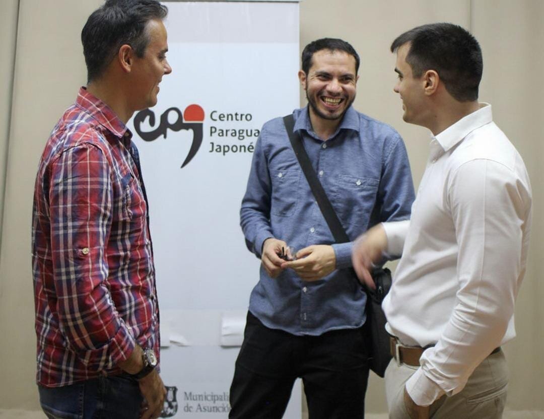 (Photo) A photograph showing three men standing indoors, talking and laughing, with a white banner displaying the logo and name of the Centro Paraguayo Japonés behind them. Text: Centro Paraguayo Japoné Municipal de Asunci[unreadable] People (three adult males) | indoor (event space or meeting room) | Three men in casual attire engaged in conversation; one man is laughing; a promotional backdrop/banner is visible. | Candid/Event photography | Colors: #FFFFFF, #A83232, #46698C, #D9C9B4 Note: The image is a realistic capture of people interacting in an indoor setting, typical of event or candid photography.