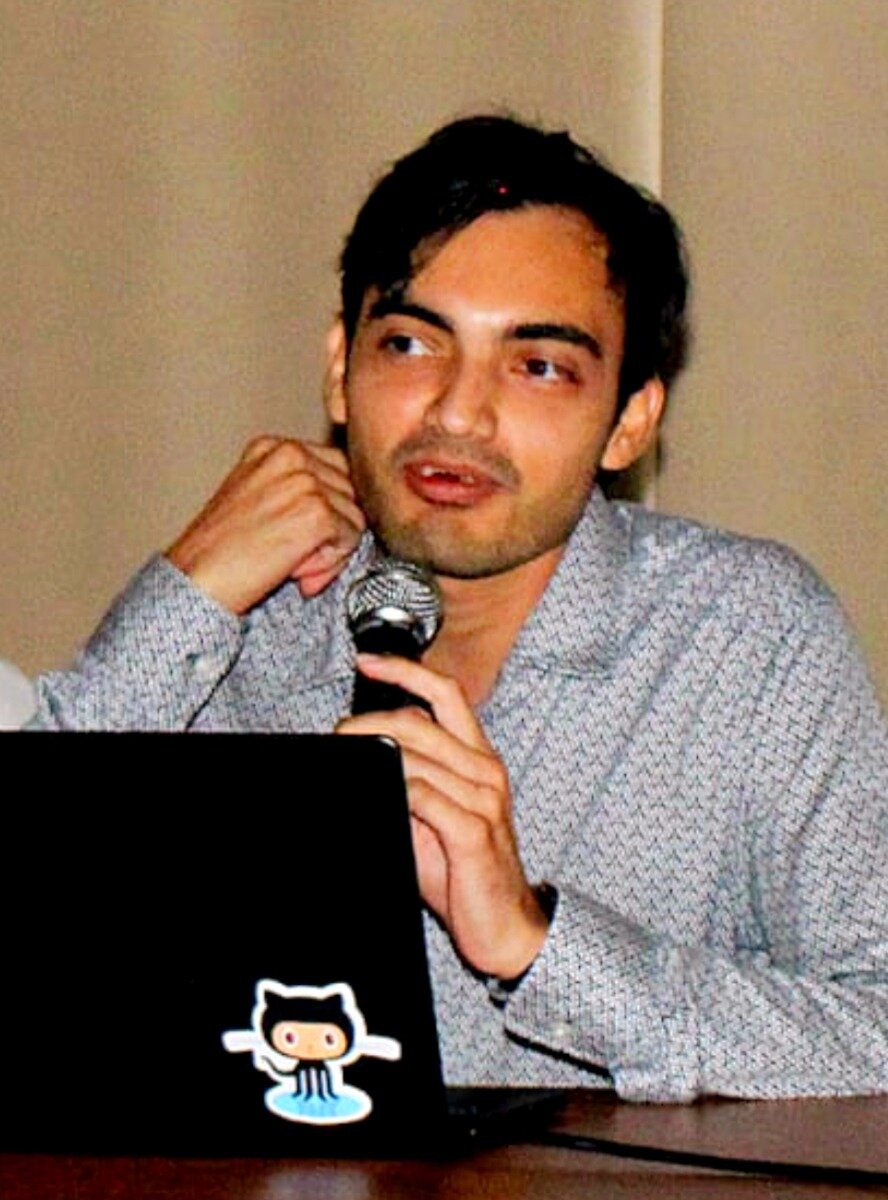 (Photo) This is a photo of a man speaking into a microphone, likely at a conference or presentation, while resting his hand on a laptop that features a GitHub Octocat sticker. person (male speaker) | indoor (likely a conference room or stage) | Holding a microphone, laptop visible with a GitHub Octocat sticker, patterned shirt. | Candid, medium close-up, slightly high contrast | Colors: #b5a48d, #000000, #d0d0d0, #c29471 Note: The image captures a real person in a specific setting (a presentation or talk), characteristic of a photograph rather than an illustration or logo.
