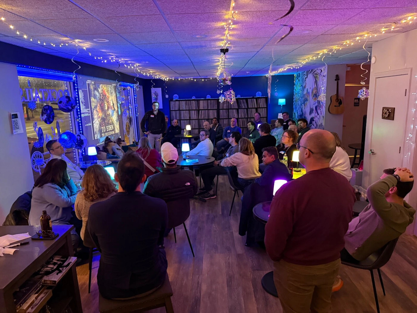 (Photo) A group of people are gathered in a dimly lit indoor space, likely a lounge or bar, watching a presentation or film projected onto a screen. The room is decorated with festive string lights and has a cozy, social atmosphere. Text: TO EDEN TO FREEDOM COCKTAILS A group of people attending a presentation or screening. | Indoor lounge or bar area. | Projection screen, string lights, multiple seated and standing individuals, guitar on the wall. | Candid event photography with ambient lighting. | Colors: #4B0082, #FFD700, #2F4F4F, #8B4513 Note: The image is a real-life photograph capturing a social event with people and a physical environment.