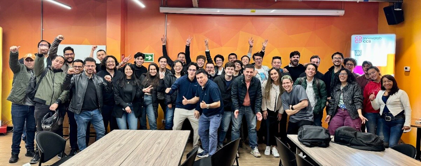 (Photo) A large group of approximately forty people is posing indoors for a group photo, standing in front of an orange patterned wall and expressing enthusiasm with raised hands and gestures. Text: Innovalab CCB CCB large group of people (team, workshop attendees) | indoor (conference room or event space) | Many subjects are smiling and making enthusiastic gestures; the background wall is a vibrant orange with a geometric pattern; a branded sign is visible on the right. | candid group portrait | Colors: #F57C00, #333333, #FFFFFF Note: This is a captured moment of real people in a real indoor setting, characteristic of a standard photograph taken at an event or workplace.
