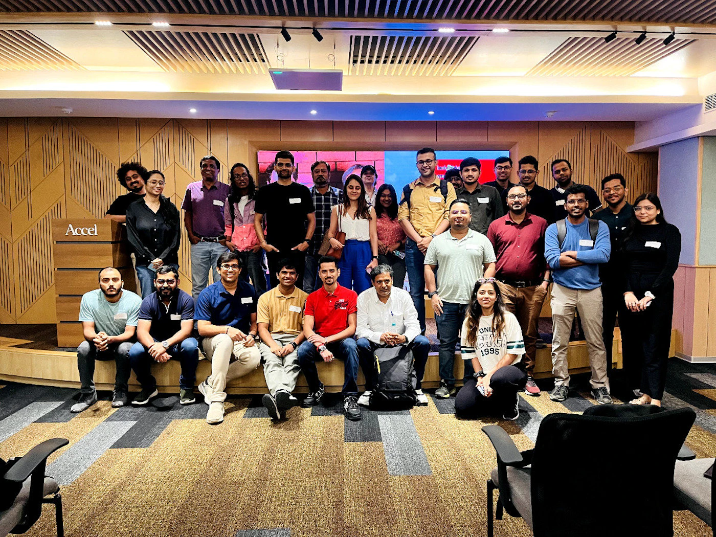 (Photo) A group of people are posing for a photo in a presentation room.  They are dressed in casual attire and appear to be at a business or professional event. Text: Accel ONLY EVER 1995 people | indoor | group photo, presentation room | group portrait Note: The image is a real-life captured moment of a group of people, clearly a photograph, not a digitally created image or logo.