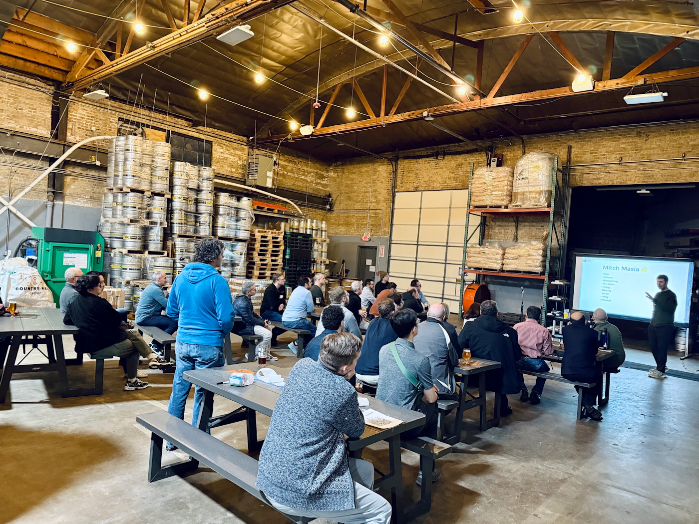 (Photo) A group of people sitting at picnic tables in a large brewery warehouse, watching a presentation given by a man standing next to a screen. Text: Mitch Masia Data Software Engineering Operations Product Strategy Marketing Sales Finance People COUNTRY MALT GROUP EXIT presentation in a brewery | indoor warehouse | stacks of beer kegs, bags of grain, picnic tables, presentation screen | candid | Colors: #8B7D6B, #3C4142, #C9B081, #2B5D90 Note: The image is a captured photograph of a real-world event and environment.