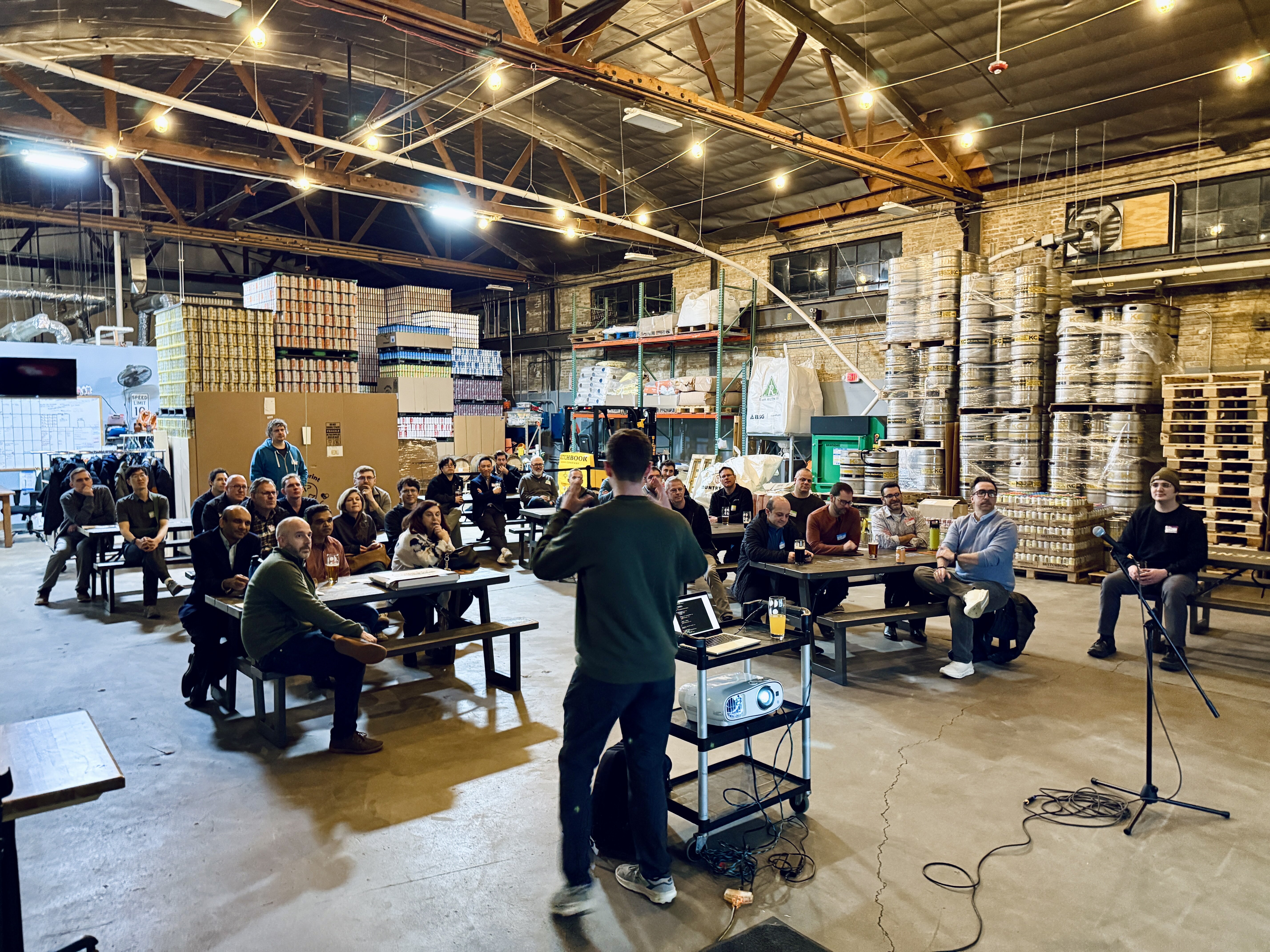 (Photo) A man stands before an audience seated at picnic tables, giving a presentation in a large warehouse or brewery filled with stacks of kegs. Text: SPEED LIMIT 10 A group of people attending a presentation in a brewery. | Indoor industrial warehouse/brewery space. | Stacks of beer kegs, audience at picnic tables, presenter with a laptop and projector. | Candid documentary photography. | Colors: #8B7D6B, #3C4142, #C5B392, #1A1C1D Note: This is a real-life photograph capturing a candid moment of a professional gathering in an industrial setting.