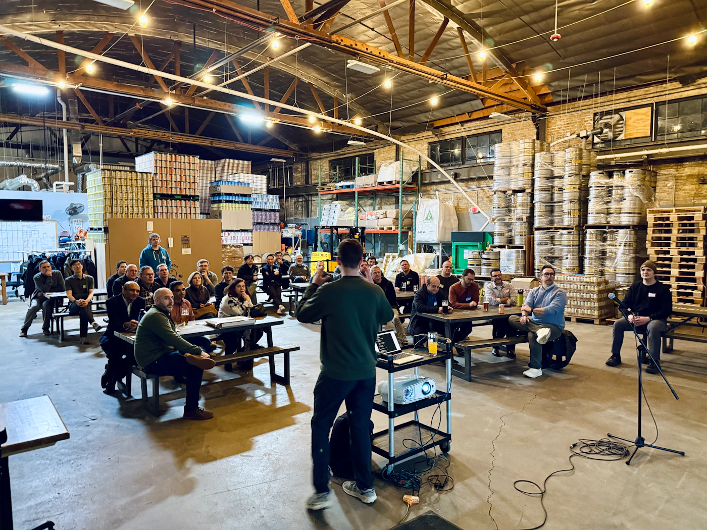 (Photo) A man stands before an audience seated at picnic tables, giving a presentation in a large warehouse or brewery filled with stacks of kegs. Text: SPEED LIMIT 10 A group of people attending a presentation in a brewery. | Indoor industrial warehouse/brewery space. | Stacks of beer kegs, audience at picnic tables, presenter with a laptop and projector. | Candid documentary photography. | Colors: #8B7D6B, #3C4142, #C5B392, #1A1C1D Note: This is a real-life photograph capturing a candid moment of a professional gathering in an industrial setting.