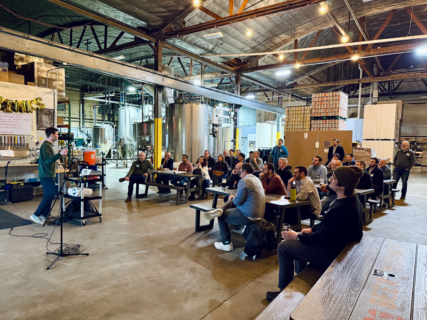 (Photo) A man stands at a podium giving a presentation to a seated audience inside a large industrial brewery with stainless steel tanks and high ceilings. Text: DRINKS presentation in a brewery | indoor, industrial | large group of people, industrial brewing equipment, speaker with microphone, gold balloons | candid, documentary | Colors: #a4a49c, #745c4c, #bcbcbc, #3c3c3c Note: This is a candid photograph capturing a real-world event in a physical environment.