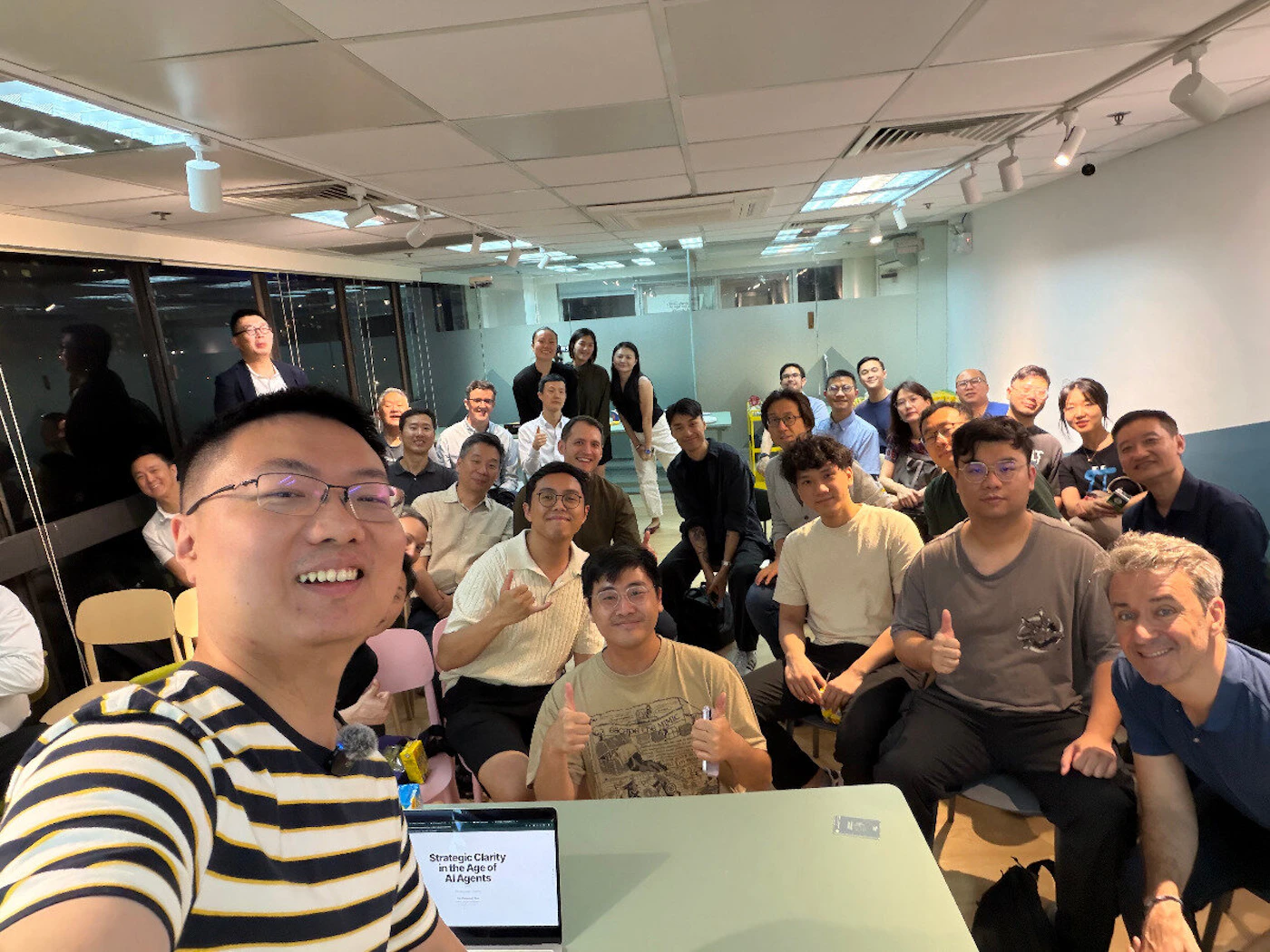 (Photo) A large group of people posing for a group selfie in an office or meeting room, with a laptop in the foreground showing a presentation slide. Text: Strategic Clarity in the Age of AI Agents group of people | indoor office | large group, selfie perspective, laptop with presentation slide | candid group portrait | Colors: #FFFFFF, #000000, #D3D3D3, #F5F5DC Note: The image is a digital photograph capturing a real-life social gathering of people in an indoor setting.