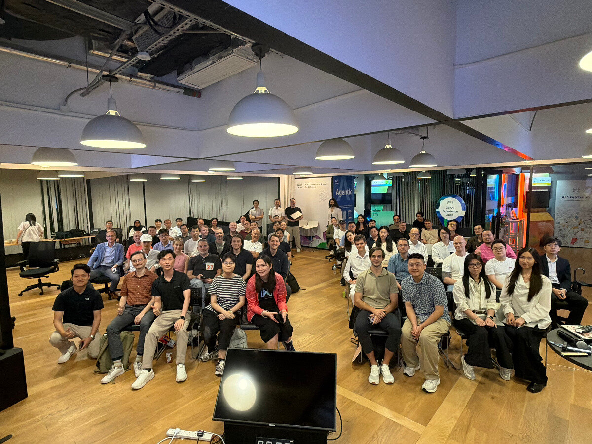 A large group of people posing for a group photo in a modern indoor event space with wooden floors and industrial-style lighting.. Text: AWS Experience Space, Agentic, GenAI Experience Zone, AWS, AWS AI Sketch Lab, 37