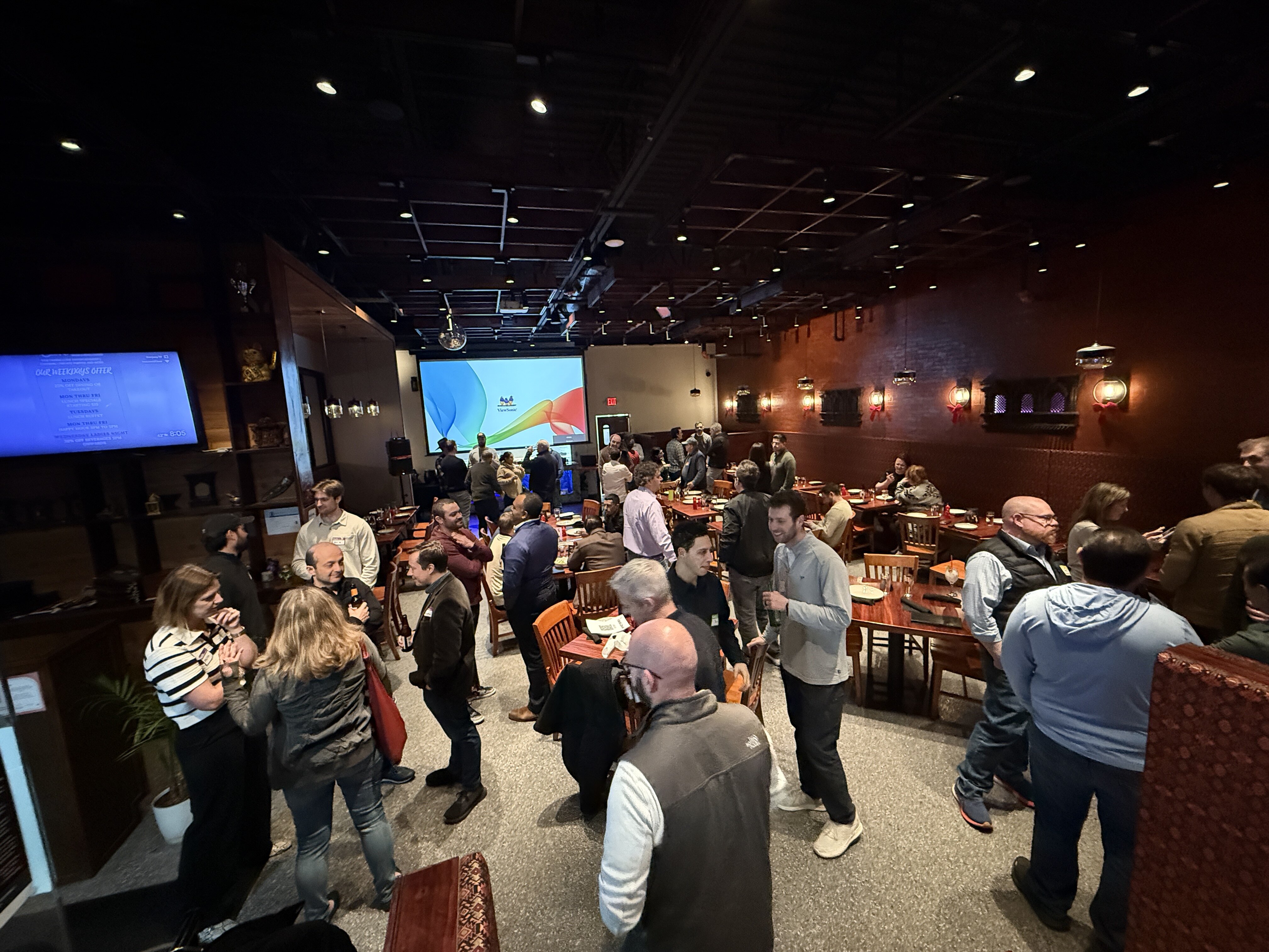 (Photo) A group of people are socializing and networking in a restaurant or event space featuring a projector screen and a TV monitor. Text: OUR BEVERAGE OFFER COCKTAILS MOCKTAILS WINE BEER SPIRITS EXIT people, social gathering | indoor, restaurant or event hall | large group, projector screen, dining tables, TV monitor | candid | Colors: #141414, #4a2c2a, #d9d9d9 Note: The image is a realistic photograph capturing a real-world scene of people at a social event.