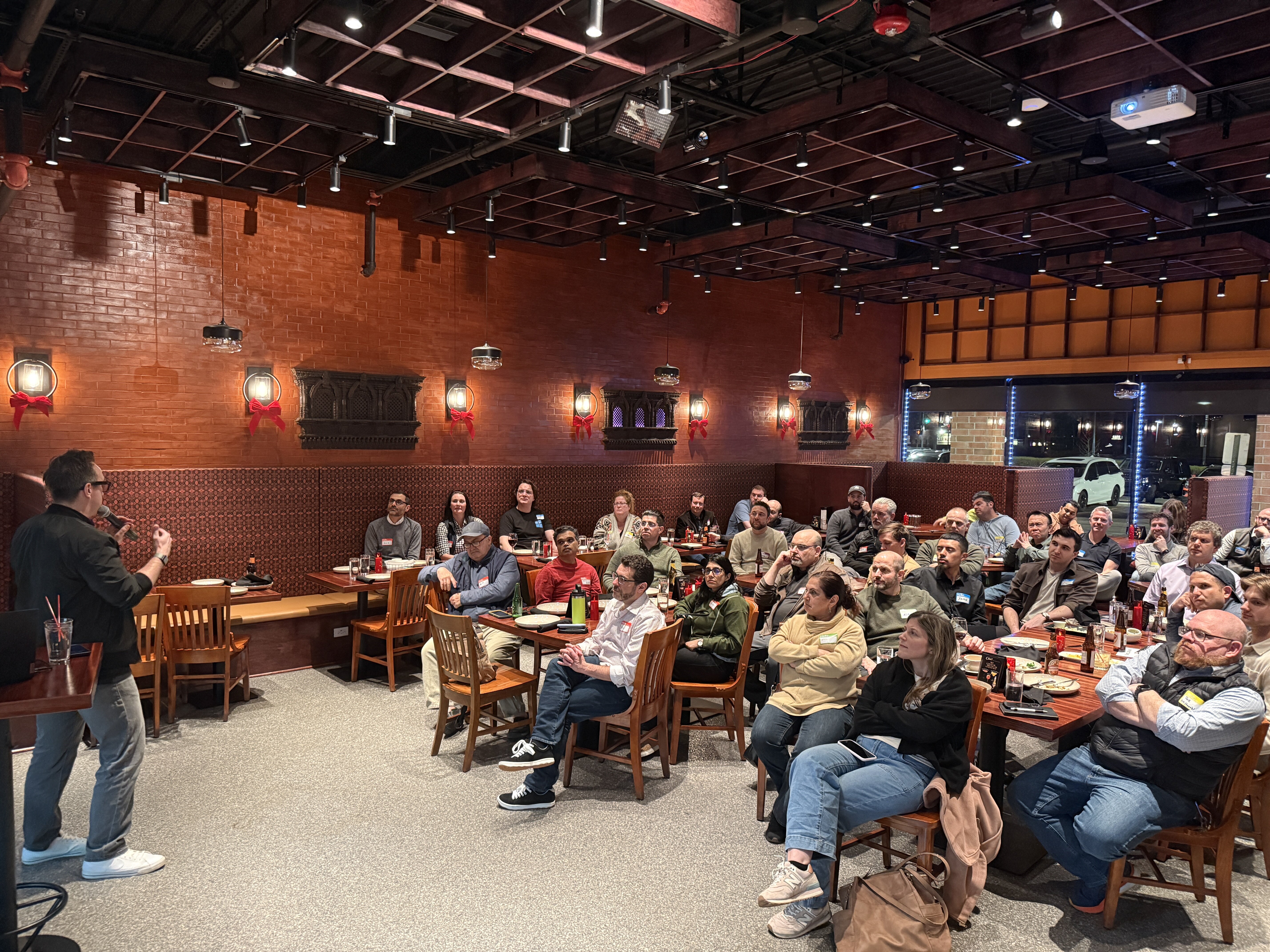 A man stands at the front of a restaurant giving a presentation to a large audience seated at tables.