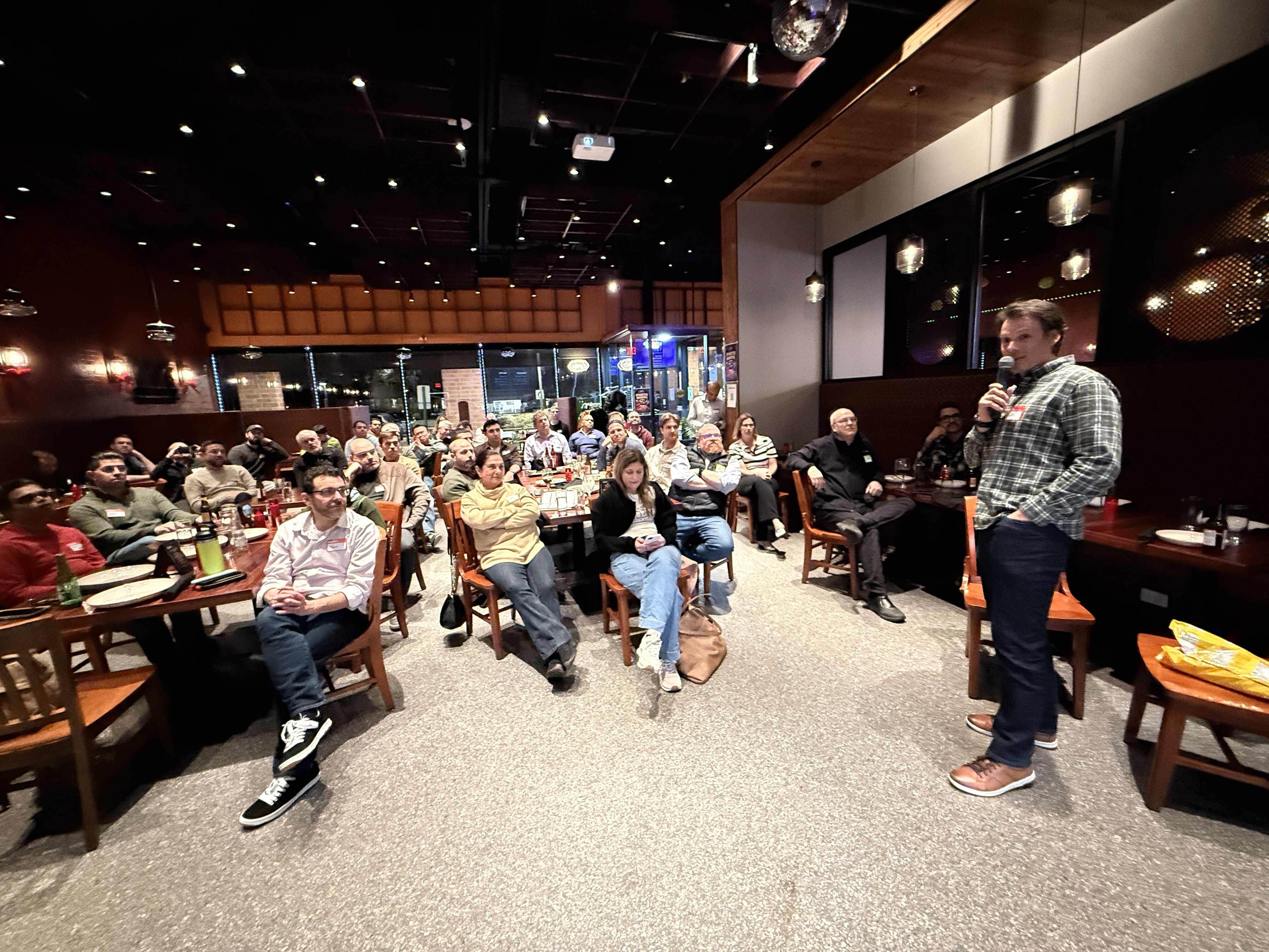 (Photo) A man in a plaid shirt speaks into a microphone to a seated audience in a dimly lit restaurant or event space. presentation | indoor | speaker with microphone, seated audience, disco ball, projector | candid | Colors: #121212, #d1cfcc, #4e3629, #5c6b5e Note: The image is a photograph capturing a real-life event with people and a physical environment.