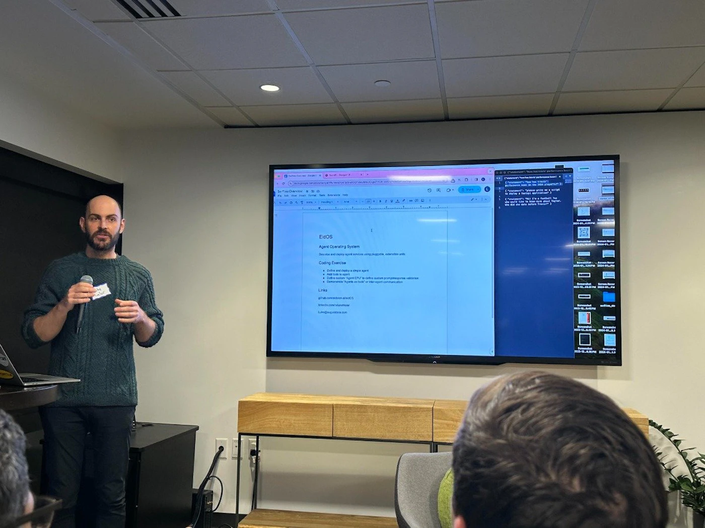 (Photo) A man is presenting in front of a large screen displaying a document and code. He's holding a microphone and addressing an audience. Text: ExOS Agent Operating System Server architecture diagram Coding Exercise 1. Define an agent. 2. Add a trait to an agent. 3. Give an example of a multi-agent conversation. 4. Create an agent that acts upon receiving a specific message. Links github.com/liquidmind/exos  person | indoor | presentation, screen with code and text, audience | candid Note: The image is a real-life captured scene of a presentation, making it a photograph.  It's not a staged image or other graphical representation.