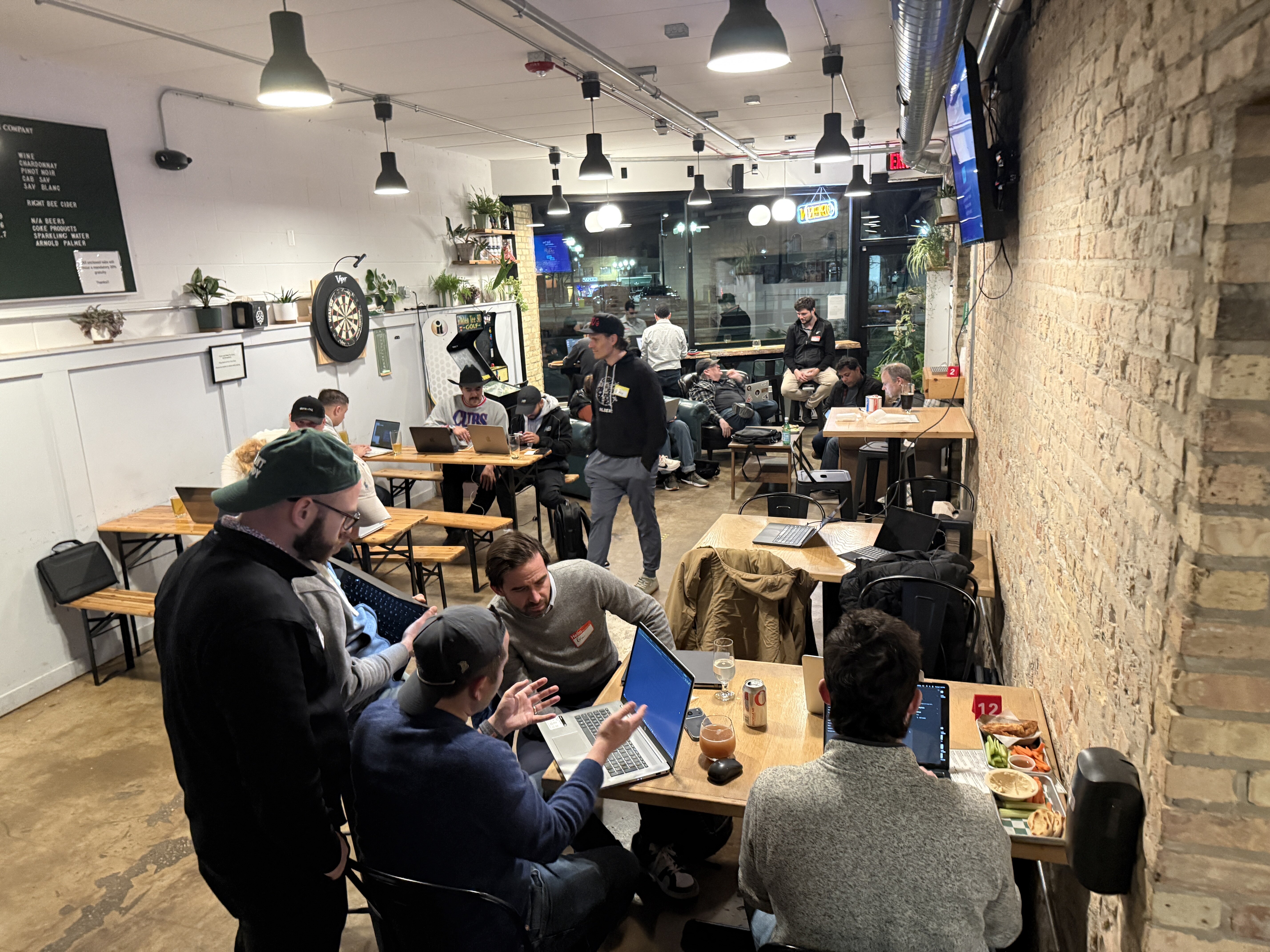 (Photo) A group of people are gathered in a cafe or bar-like setting, many of whom are using laptops and engaged in discussions, suggesting a tech meetup or co-working event. Text: WINE CHARDONNAY PINOT NOIR CAB SAV SAUV BLANC EIGHT BEE BEER MIX BEERS SODA PRODUCTS SPARKLING WATER ARNOLD PALMER OPEN 12 CITIES people | indoor | Multiple people with laptops in a social, tech-oriented atmosphere. | candid | Colors: #E6E1D3, #3B3B3B, #8B4513 Note: The image is a real-life photograph capturing a candid scene of people in an indoor environment.
