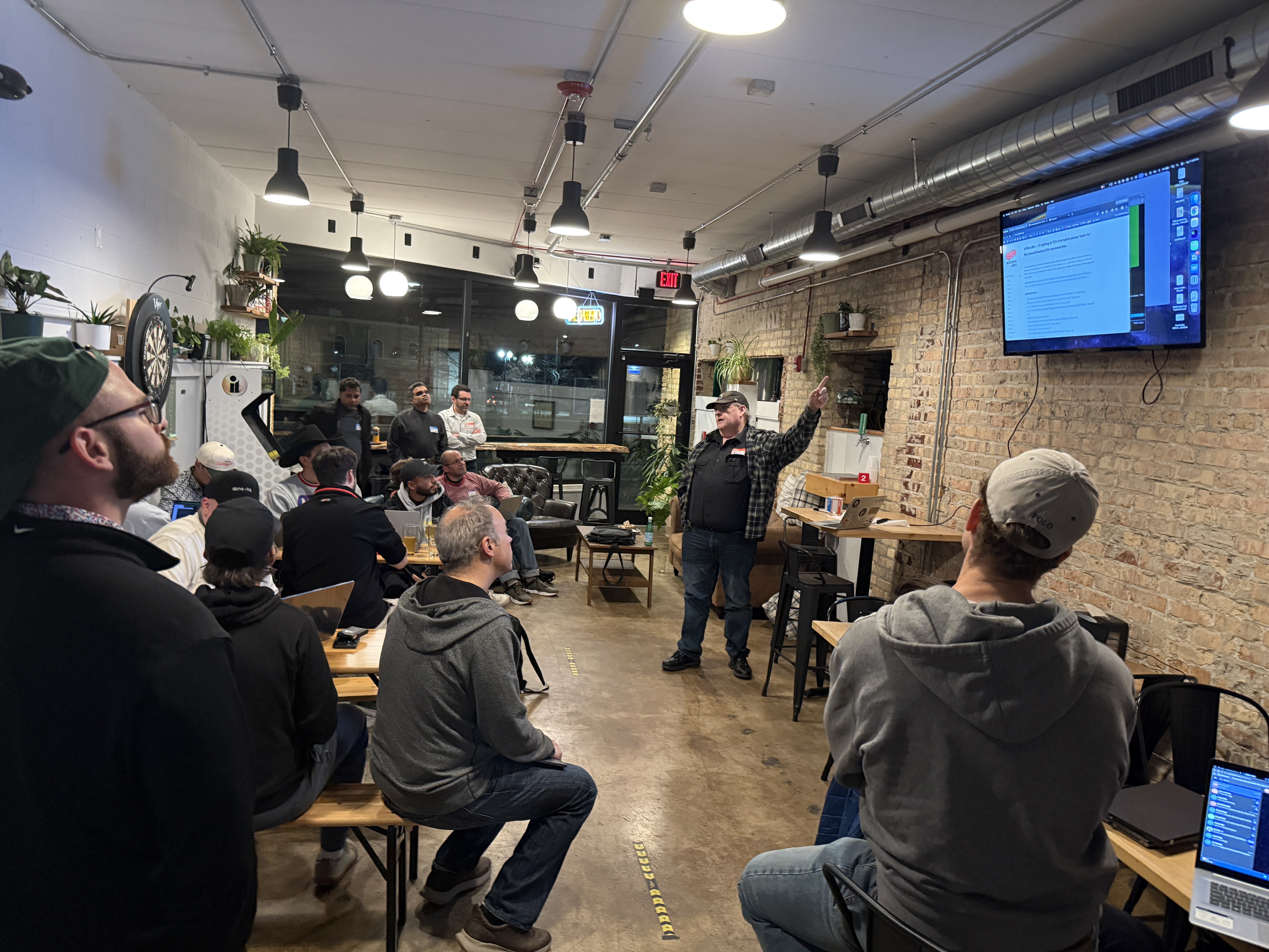 (Photo) A group of people are gathered in an indoor space with brick walls, watching a presentation on a wall-mounted TV screen. A man stands at the front, pointing toward the screen as he speaks to the audience. Text: EXIT, Documentation, Getting Started, Introduction, 2 people, indoor presentation | indoor, bar or cafe | brick walls, wall-mounted TV, group of men, presenter pointing, laptops | candid | Colors: #8b7d6b, #2c2c2c, #f5f5f5, #a6937c Note: This is a candid photograph capturing a real-world event with people in an indoor environment.