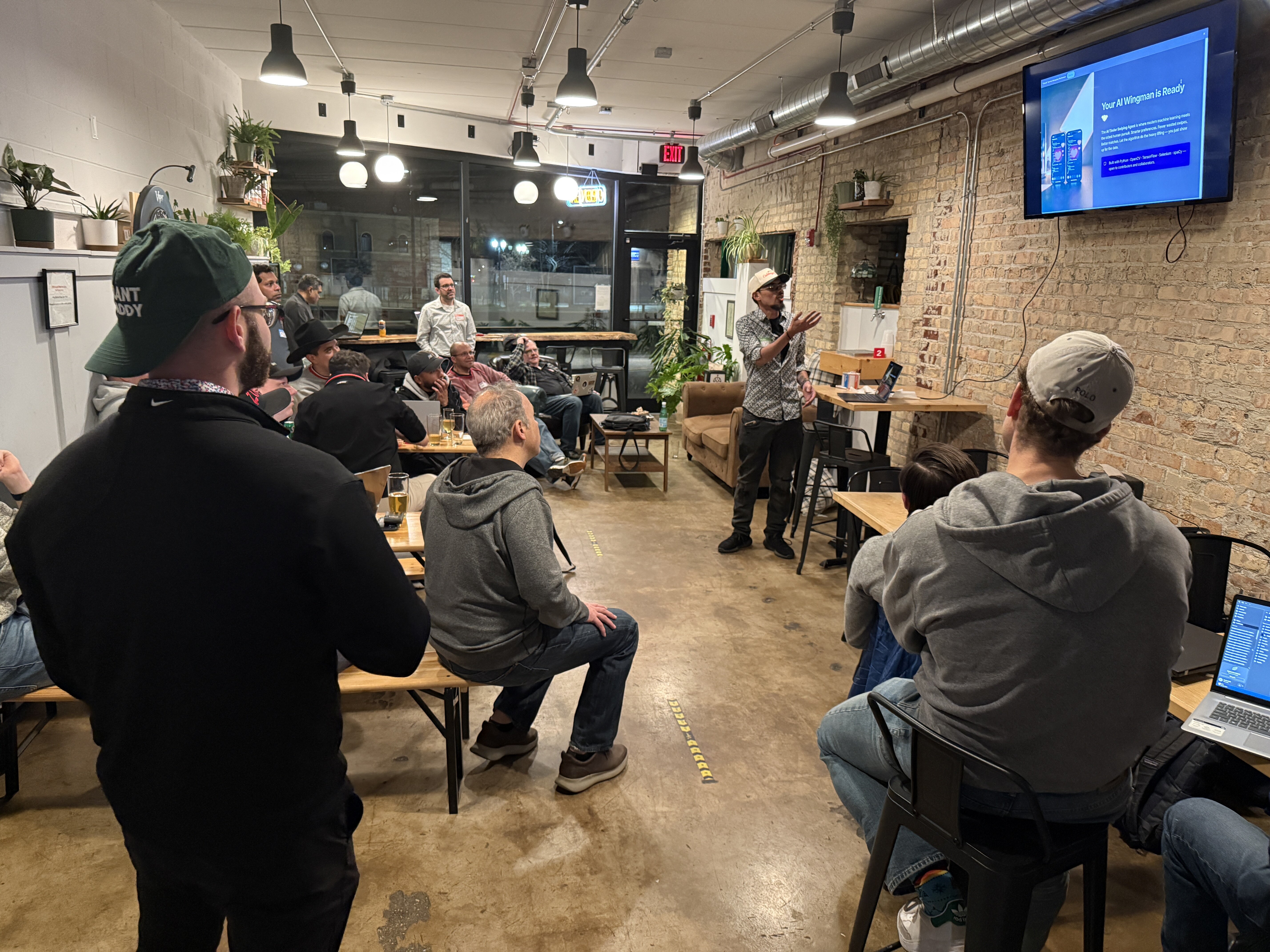 (Photo) A man gives a presentation about an 'AI Wingman' app to a small audience in a room with exposed brick walls and a casual atmosphere. Text: Your AI Wingman is Ready PLANT DADDY EXIT people at a presentation | indoor, likely a cafe, bar, or co-working space | exposed brick wall, wall-mounted TV screen, audience members with laptops, casual attire | candid | Colors: #8B735B, #2B2B2B, #E5E5E5 Note: The image is a captured photograph of a real-world event featuring people, an indoor setting, and physical objects.