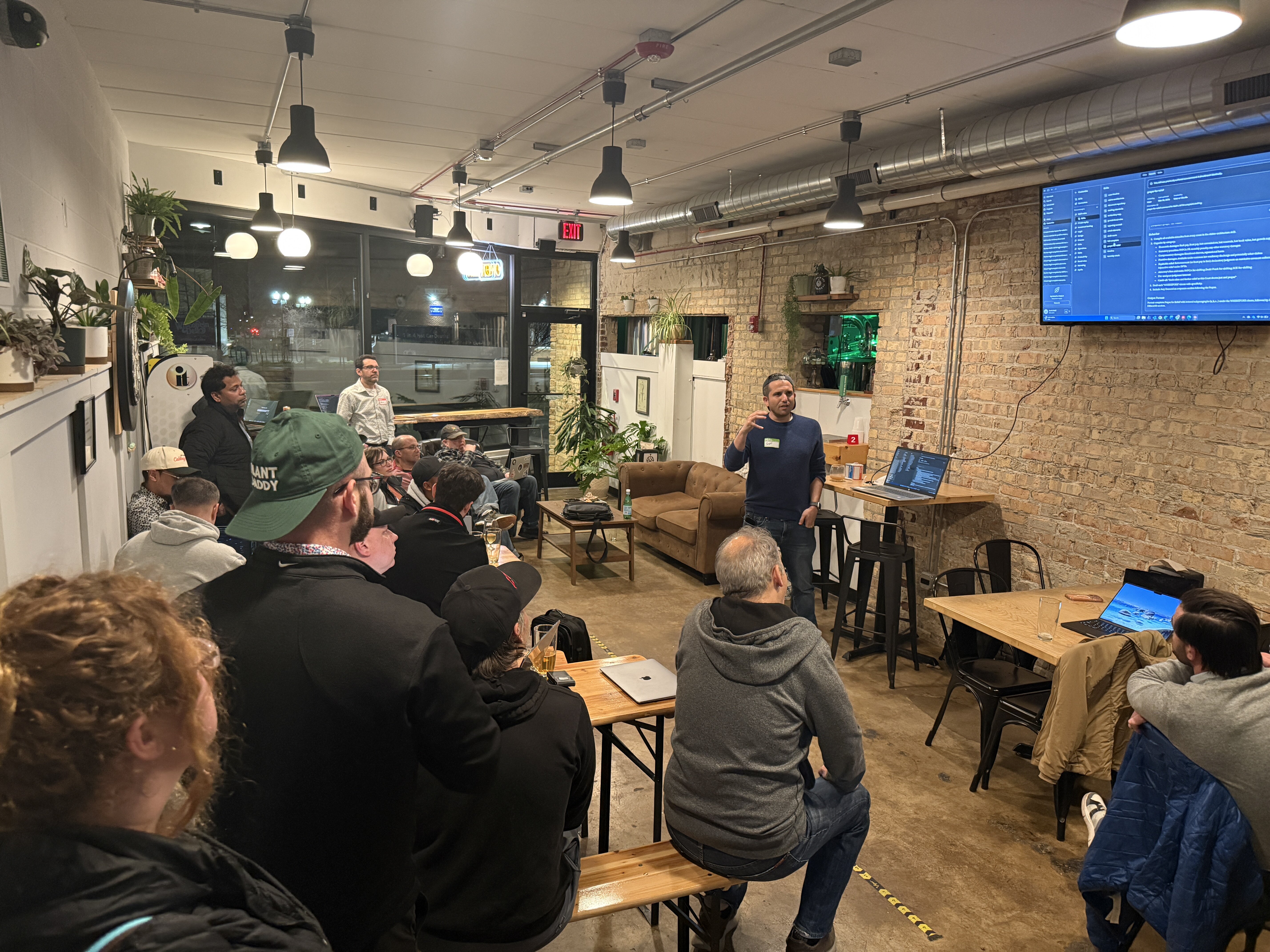 (Photo) A man is giving a presentation to a group of people in an indoor space with exposed brick walls, while a large screen displays code. Text: PLANT DADDY, EXIT presentation | indoor | speaker, audience, TV screen with code, exposed brick wall, industrial lighting | candid | Colors: #b58e6d, #2b2b2b, #968165, #dcdcdc Note: The image is a photograph capturing a real-life scene involving people, interior architecture, and electronic devices.