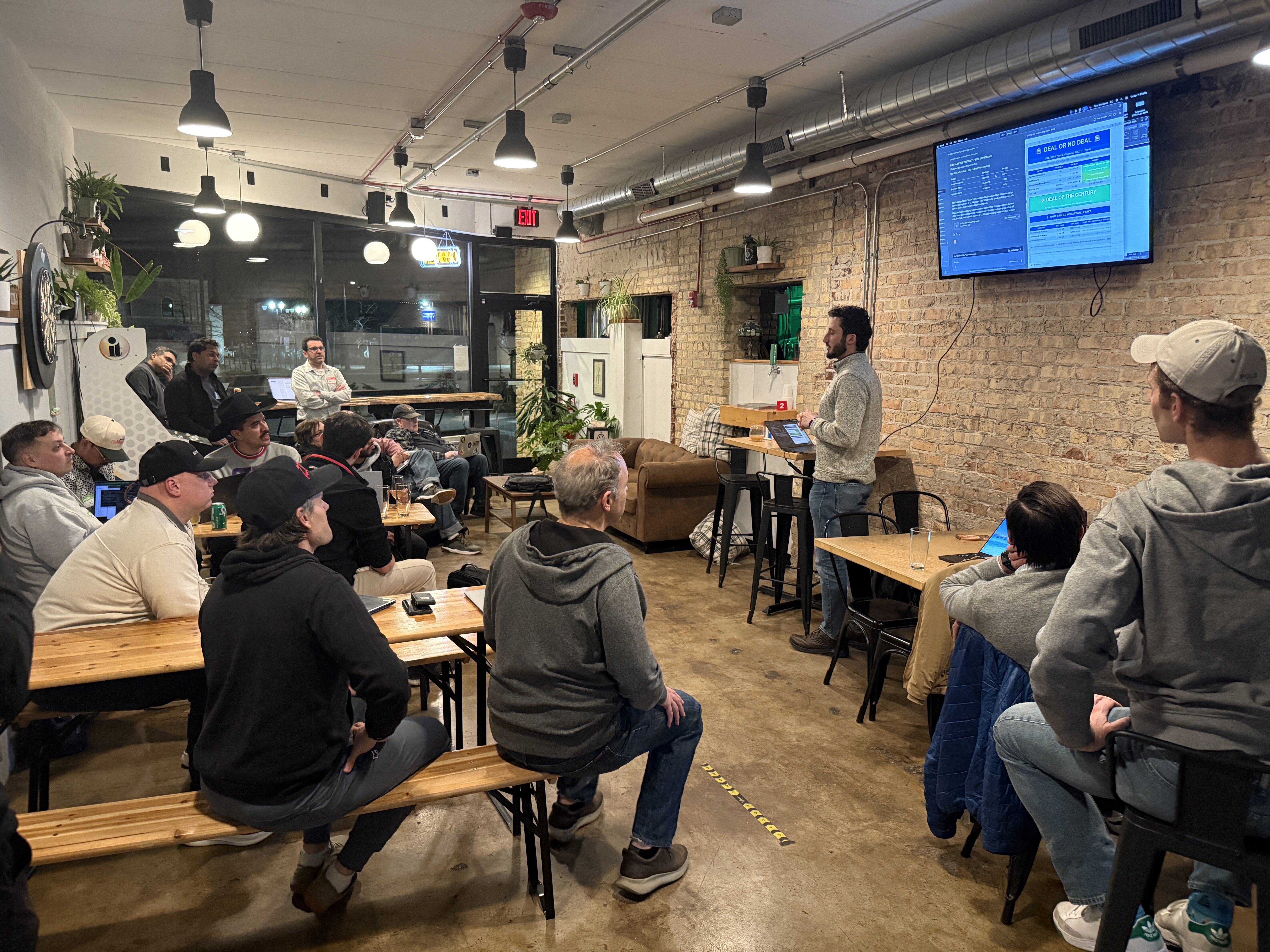 (Photo) A man is giving a presentation from a laptop to a seated audience in a room with exposed brick walls and industrial lighting. Text: DEAL OR NO DEAL, EXIT presentation | indoor | exposed brick walls, industrial ceiling pipes, audience on benches, presenter with laptop, TV screen display | candid | Colors: #8B4513, #333333, #E0E0E0 Note: The image is a real-life photograph capturing a candid scene of a presentation in an indoor environment.