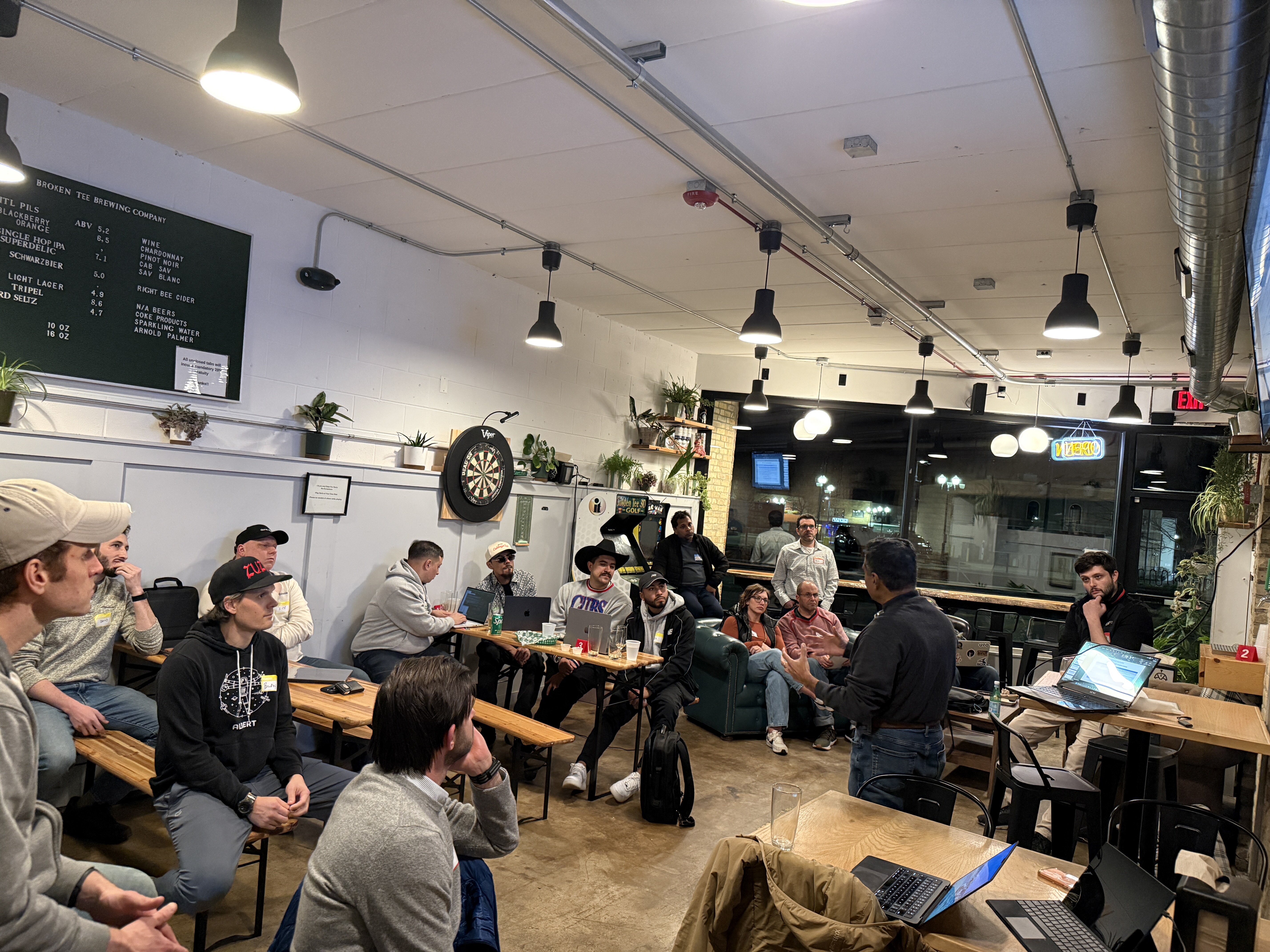 A group of people are gathered in a brewery taproom, listening to a man give a presentation. Several audience members have laptops open on tables in front of them.. Text: BROKEN TEE BREWING COMPANY, PILS, BLACKBERRY ORANGE, SINGLE HOP IPA, SUPERDELIC, SCHWARZBIER, LIGHT LAGER, TRIPEL, HARD SELTZER, ABV 5.2, 8.5, 7.1, 5.0, 4.8, 4.7, WINE, CHARDONNAY, PINOT NOIR, CAB SAUV, SAUV BLANC, RIGHT BEE CIDER, N/A BEERS, CORE PRODUCTS, SPARKLING WATER, ARNOLD PALMER, 10 OZ, 16 OZ, Viper, CITRA, EXIT, OPEN