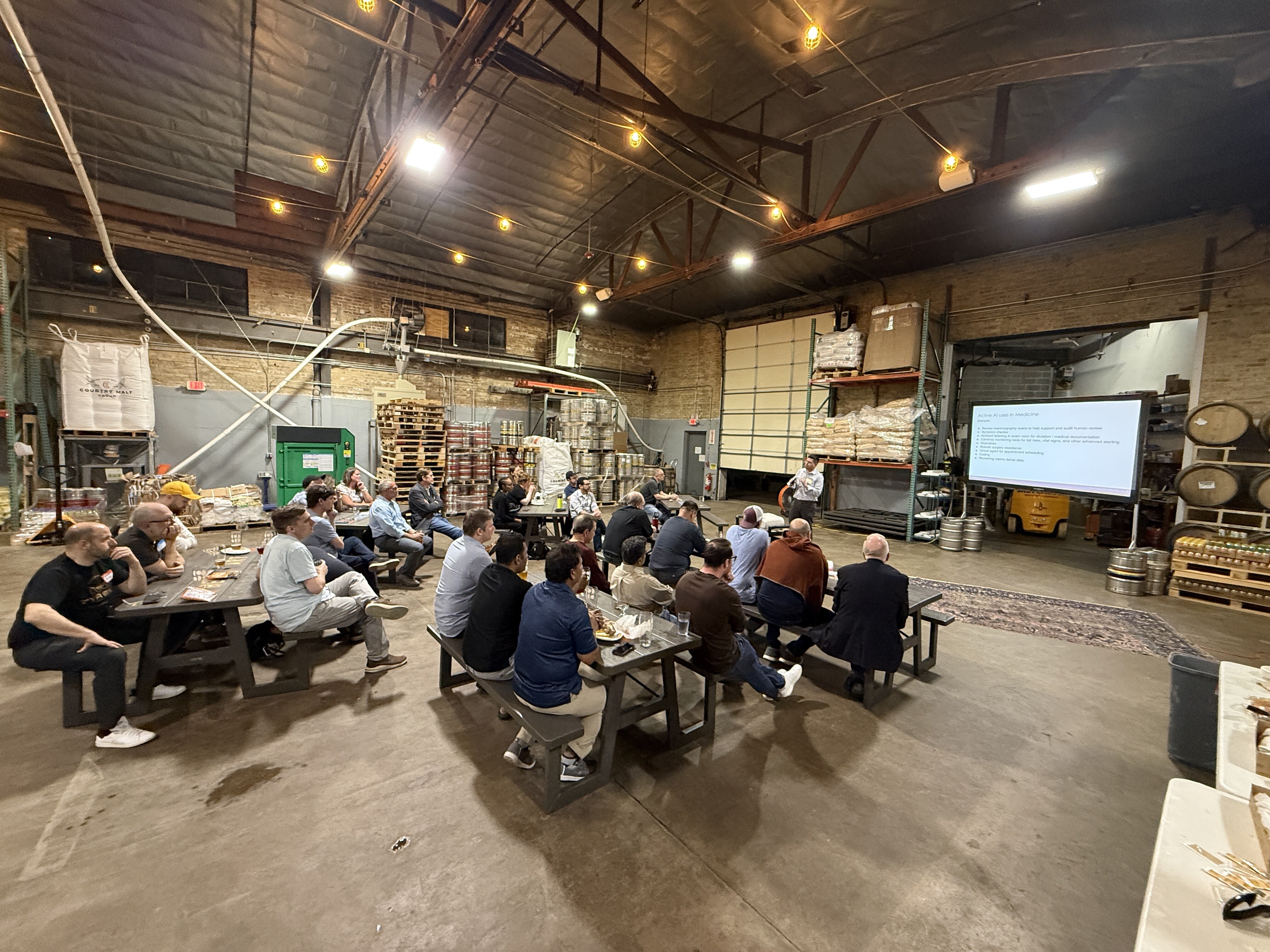 (Photo) A group of people are seated at picnic tables in a large, industrial warehouse or brewery setting, attending a presentation shown on a large projector screen. Text: Building the Future of Manufacturing Challenges: - The manufacturing workforce is aging and skilled workers are in short supply. - Global supply chain disruptions and economic uncertainty. - Legacy systems and technical debt hindering digital transformation. - Data silos and lack of interoperability across the value chain. - Cybersecurity threats and the need for robust data protection. - Sustainability and ESG requirements. CHOPPING BLOCK A business or educational presentation in an industrial setting. | Indoor, likely a brewery or warehouse with high ceilings and industrial equipment. | large projector screen | wooden barrels | string lights | picnic tables | industrial shelving | Candid wide-angle shot | Colors: #4a4a4a, #a68a64, #ffffff, #2e2e2e Note: The image is a real-life photograph capturing a scene of people in an indoor environment.