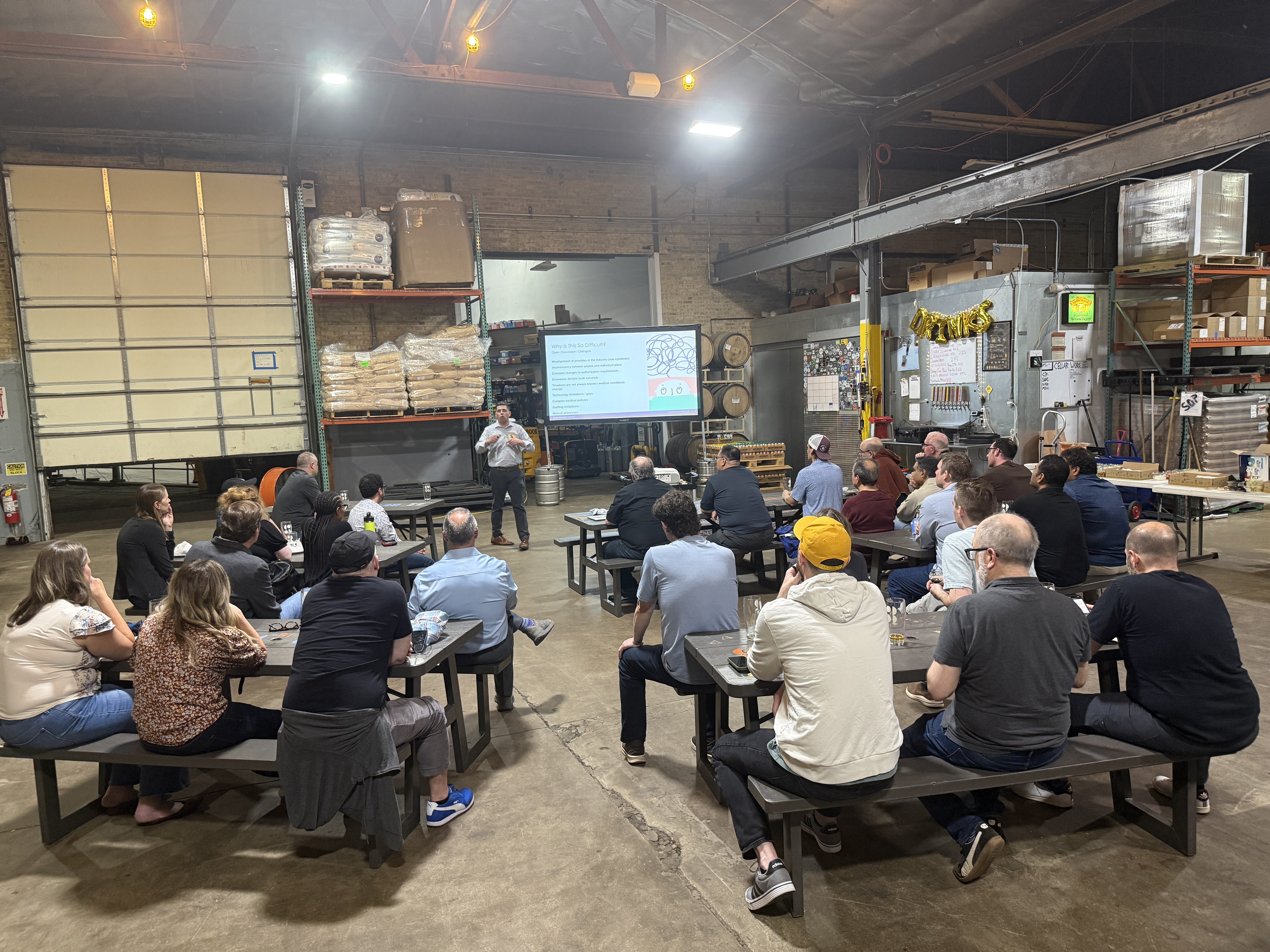 A man stands at the front of a large warehouse space giving a presentation to an audience seated at picnic tables.. Text: DRINKS