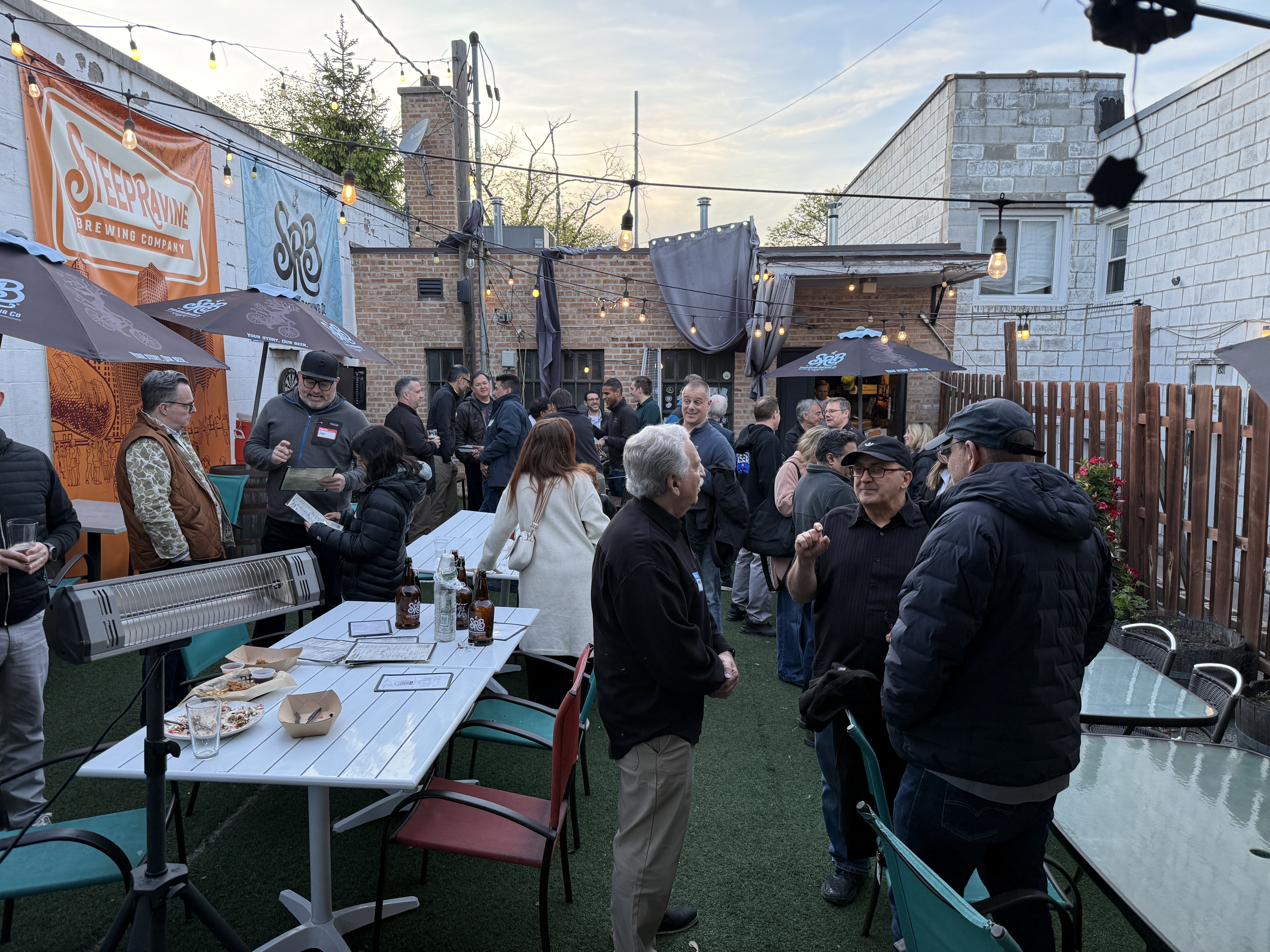 (Photo) A group of people are gathered for a social event in an outdoor brewery patio area featuring string lights and banners. Text: STEEP RAVINE BREWING COMPANY SRB social gathering at a brewery | outdoor patio | multiple people, brewery branding, string lights, patio furniture | candid | Colors: #5e6b5e, #d1603d, #f2f2f2, #2e2e2e Note: This is a real-world photograph capturing a candid scene of people in a physical environment.