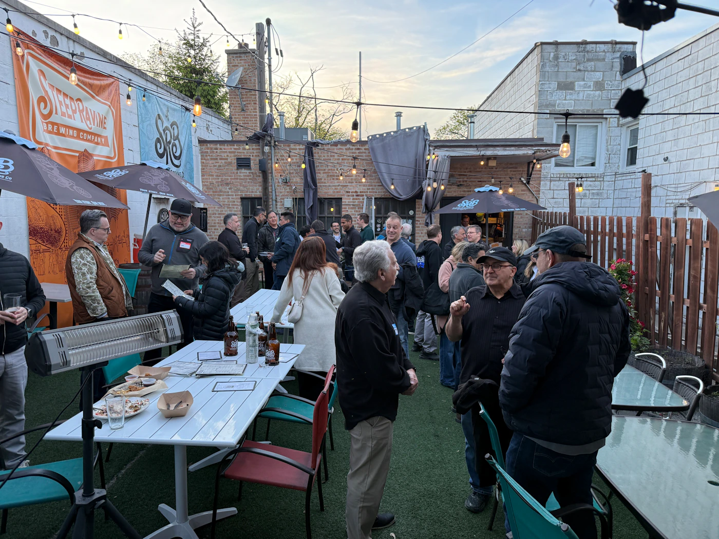 (Photo) A group of people are gathered for a social event in an outdoor brewery patio area featuring string lights and banners. Text: STEEP RAVINE BREWING COMPANY SRB social gathering at a brewery | outdoor patio | multiple people, brewery branding, string lights, patio furniture | candid | Colors: #5e6b5e, #d1603d, #f2f2f2, #2e2e2e Note: This is a real-world photograph capturing a candid scene of people in a physical environment.