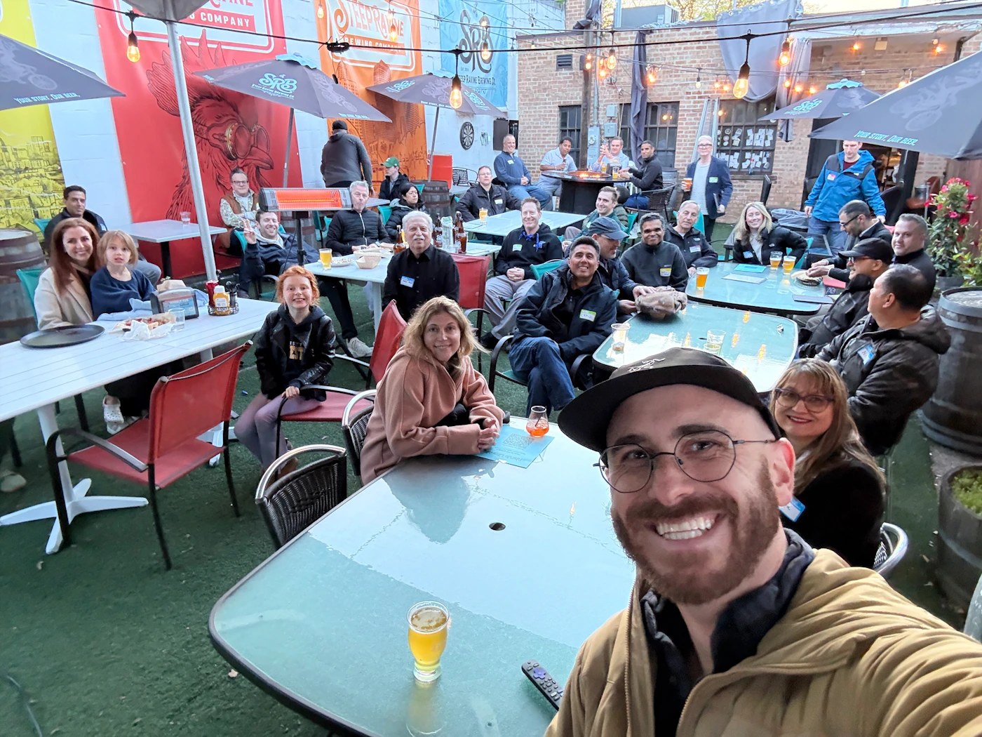 (Photo) A large group of people are socialising at an outdoor brewery patio, with several seated at tables and a man in the foreground taking a selfie. Text: STEEPLEJACK BREWING COMPANY, SRB, BEER WINE SPIRITS, YOUR STORY, OUR BEER group of people at an outdoor patio | outdoor patio, brewery/restaurant | multiple people, tables, drinks, banners, string lights, artificial turf | candid, selfie | Colors: #D0312D, #3B719F, #4B6A3A, #FFFFFF, #000000 Note: The image is a real-life photograph capturing a candid social moment at an outdoor venue.