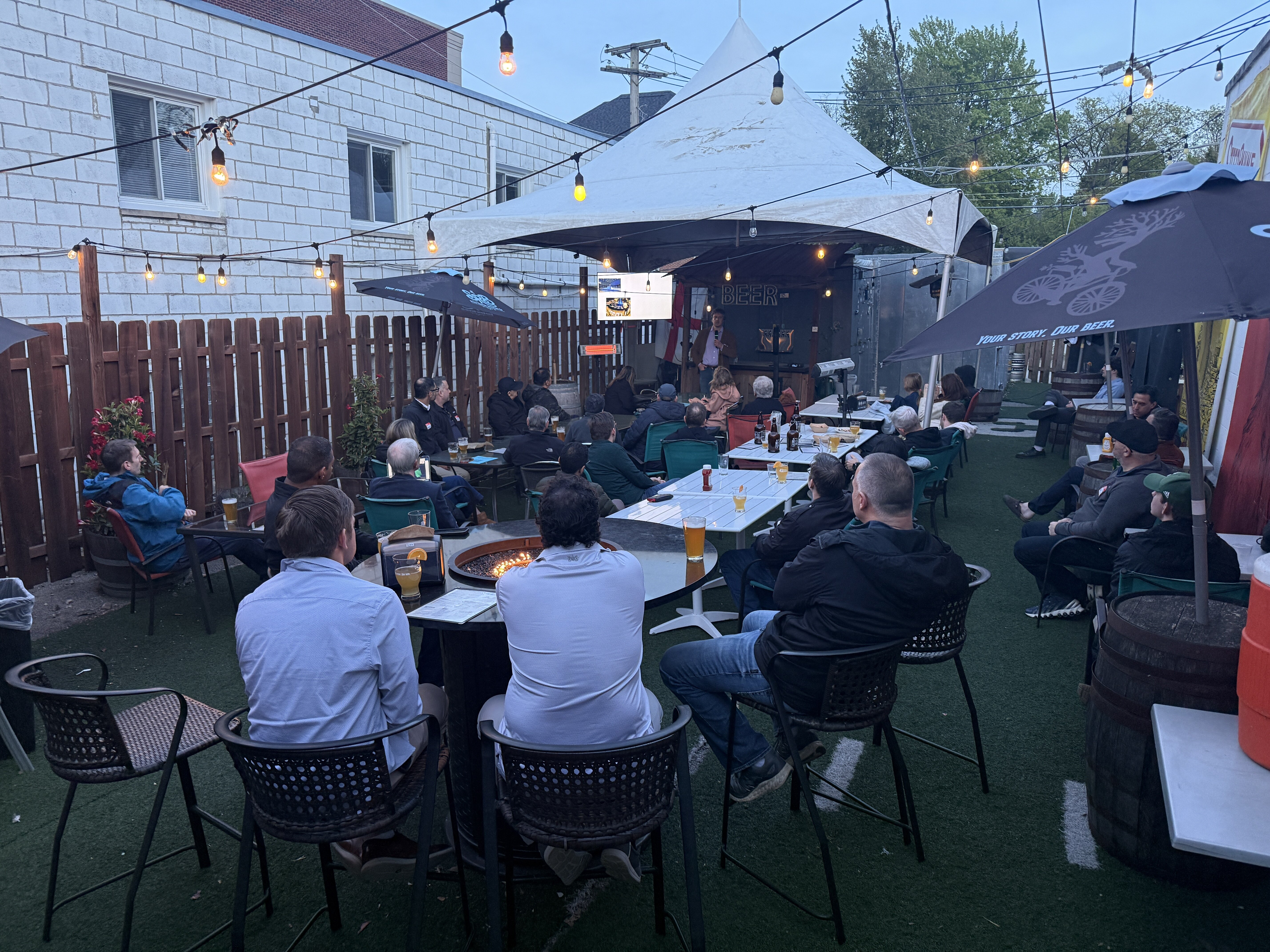 (Photo) A group of people are gathered at an outdoor patio at dusk, many sitting at tables and watching a presentation being given under a white tent. Text: BEER YOUR STORY. OUR BEER. A group of people attending an outdoor presentation. | Outdoor beer garden or patio at dusk. | String lights, fire pit, presentation screen, white tent, artificial turf. | Candid wide-angle shot. | Colors: #1a2a1a, #7b5e43, #d9d9d9, #3a4a5a Note: This is a real-life photograph capturing a candid scene of people at an outdoor social and professional event.