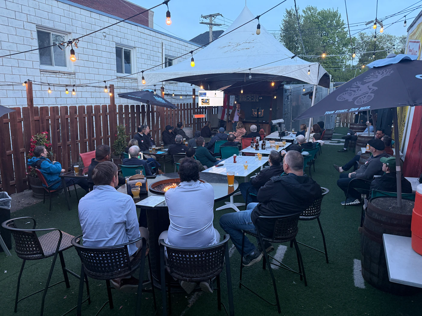 (Photo) A group of people are gathered at an outdoor patio at dusk, many sitting at tables and watching a presentation being given under a white tent. Text: BEER YOUR STORY. OUR BEER. A group of people attending an outdoor presentation. | Outdoor beer garden or patio at dusk. | String lights, fire pit, presentation screen, white tent, artificial turf. | Candid wide-angle shot. | Colors: #1a2a1a, #7b5e43, #d9d9d9, #3a4a5a Note: This is a real-life photograph capturing a candid scene of people at an outdoor social and professional event.
