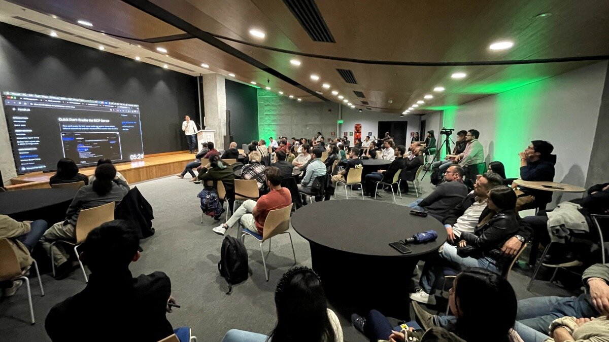 The image shows a presentation in a large room with an audience. A person is standing on a stage presenting something displayed on a large screen.. Text: Quick Start Guide to HCP Server
05:24