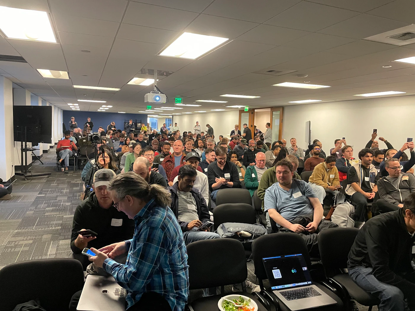 (Photo) A large group of people are seated in rows of chairs in a modern office or conference room, many of whom are looking at their phones or laptops. Text: EXIT 60 Large group of people | Indoor office or conference room | Rows of black chairs, ceiling-mounted projector, multiple people using mobile devices and laptops. | Candid wide-angle | Colors: #f0f0f0, #2c2c2c, #5a5a5a, #3b5998 Note: The image is a realistic photograph capturing a real-world indoor scene with people.