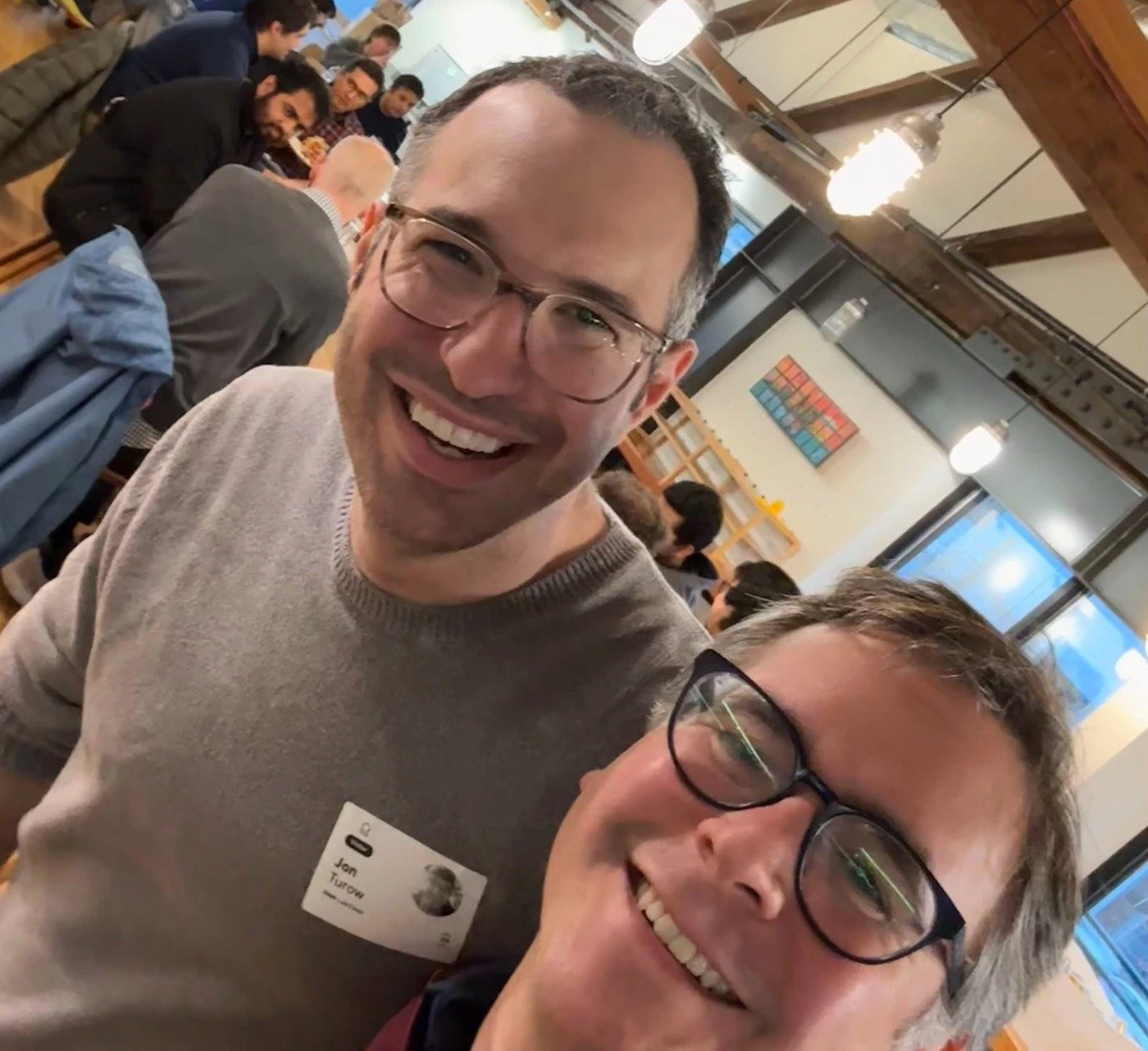 (Photo) A selfie of two smiling men wearing glasses, taken in a busy indoor space.  One of the men is wearing a name tag. Text: Jan Turow person | indoor | two people, name tag, indoor setting, other people in the background | selfie Note: This image is a photograph capturing a moment in time with real people in a real-world setting.