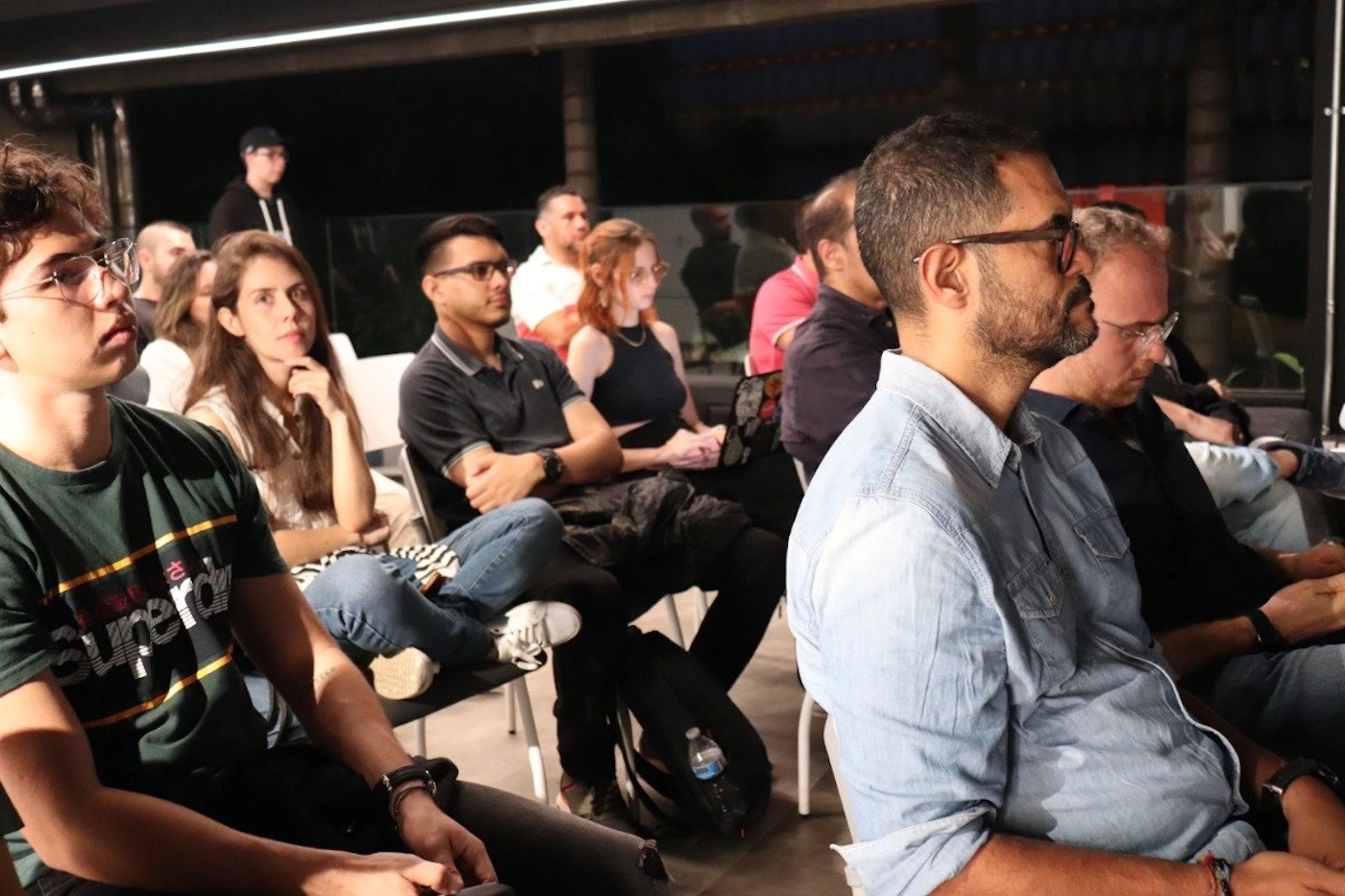 (Photo) A photograph of a group of people sitting in chairs, appearing to be part of an audience or attending a presentation. Text: Superdry people | indoor | group of people, audience setting | candid Note: The image is a real-life captured scene of people in a setting, indicative of a photograph rather than a digitally created image or graphic design.