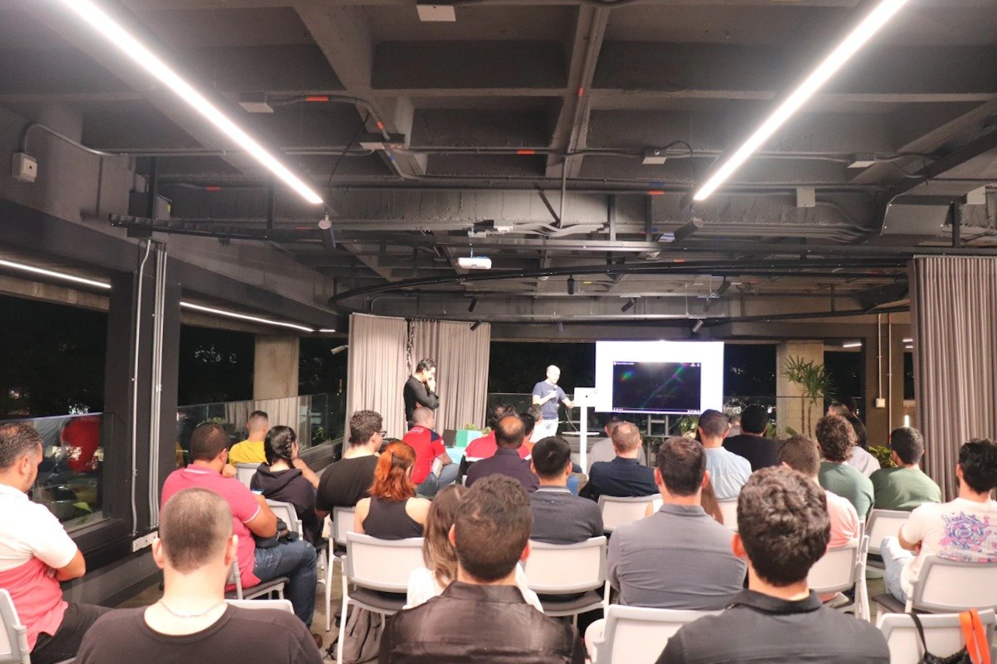 (Photo) A photo of a presentation taking place in a modern, dimly lit room. An audience sits in chairs facing a presenter and a screen displaying a graphic. people | indoor | presentation, audience, screen | candid Note: The image is a real-life captured image of a scene, depicting a presentation with an audience and a speaker.  It exhibits characteristics of a photograph, such as realistic lighting and perspective.