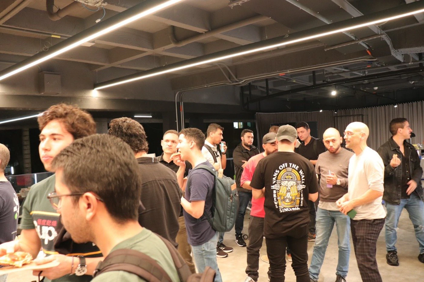 (Photo) A group of people are gathered in a dimly lit indoor space, seemingly socializing and enjoying refreshments. Text: SCALE UP VANS OFF THE WALL SINCE 1966 people | indoor | group of people, casual setting, food and drinks | candid Note: The image is a candid photograph capturing a real-life scene of a gathering. It depicts people in a casual setting, likely an event or social function.
