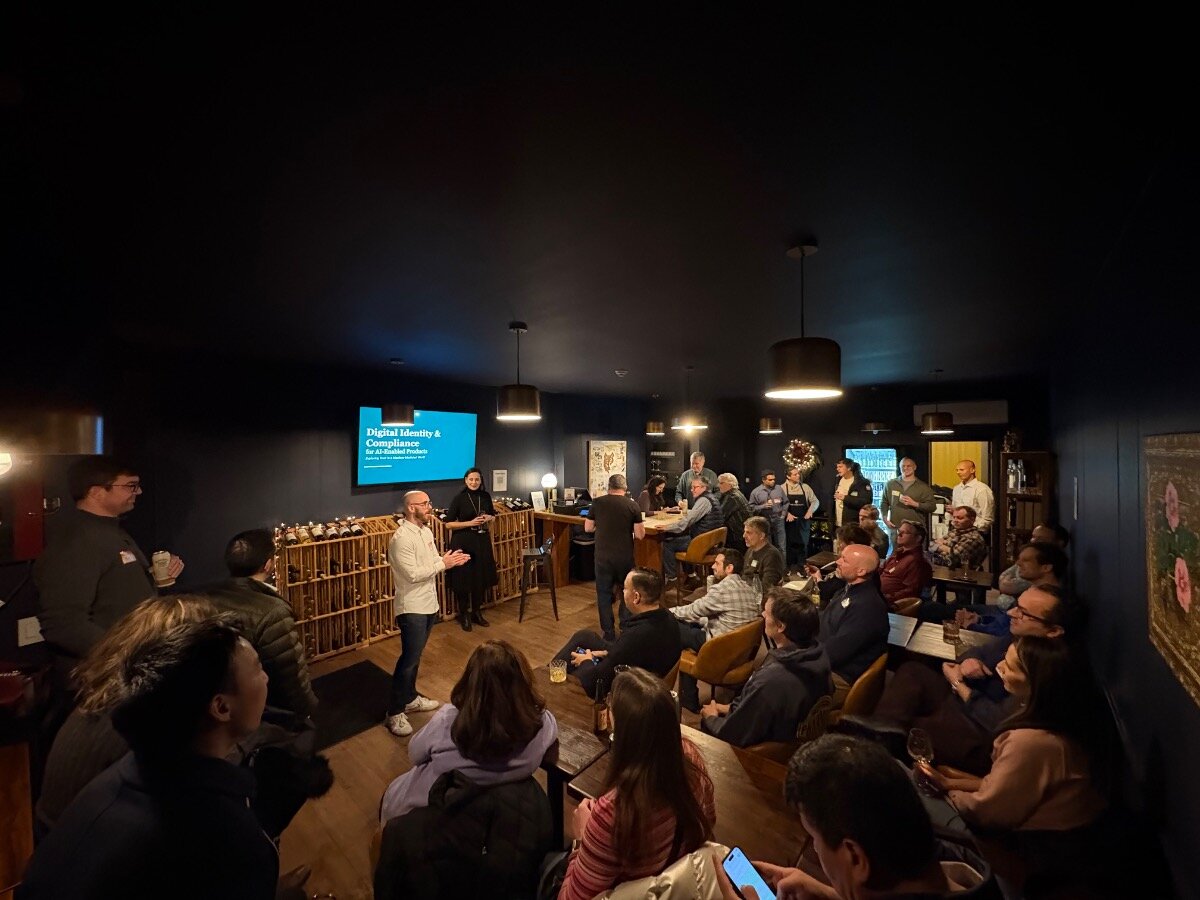 (Photo) A photograph captures a technical presentation or meetup taking place in a dimly lit, intimate indoor venue with dark blue walls, showing a speaker addressing a large audience gathered around tables. Text: Digital Identity & Compliance for AI-Related Products Group of people attending a presentation/meetup | Indoor, dark lounge or private event room | Speaker, presentation screen, large audience, wine rack storage, low lighting | Candid, event documentation | Colors: #192333, #0A0A0A, #FFFFFF, #A8855F Note: This is a photograph capturing a real-world scene of an indoor event, specifically a presentation or discussion group.