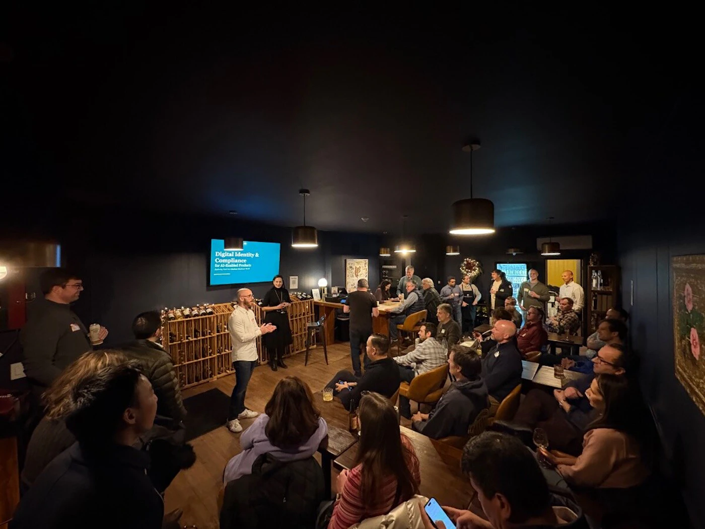 (Photo) A photograph captures a technical presentation or meetup taking place in a dimly lit, intimate indoor venue with dark blue walls, showing a speaker addressing a large audience gathered around tables. Text: Digital Identity & Compliance for AI-Related Products Group of people attending a presentation/meetup | Indoor, dark lounge or private event room | Speaker, presentation screen, large audience, wine rack storage, low lighting | Candid, event documentation | Colors: #192333, #0A0A0A, #FFFFFF, #A8855F Note: This is a photograph capturing a real-world scene of an indoor event, specifically a presentation or discussion group.