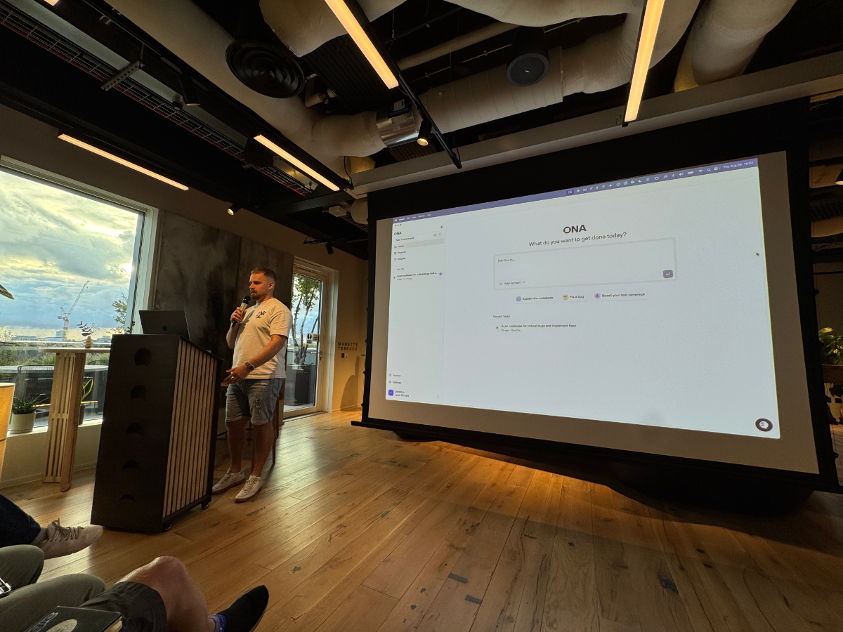 A photograph captures a man giving a presentation in a modern room with large windows, addressing an unseen audience. A large projection screen behind him displays a task management interface with text prompts.. Text: ONA
What do you want to get done today?
Just like that...
+ Add new item
Rephrase the question
Fix a typo
Boost your text with AI
Recent tasks
Run automated tests to critical bug for new implement feature
Thu Apr 25, 14:04
MANETTI SHREVE
