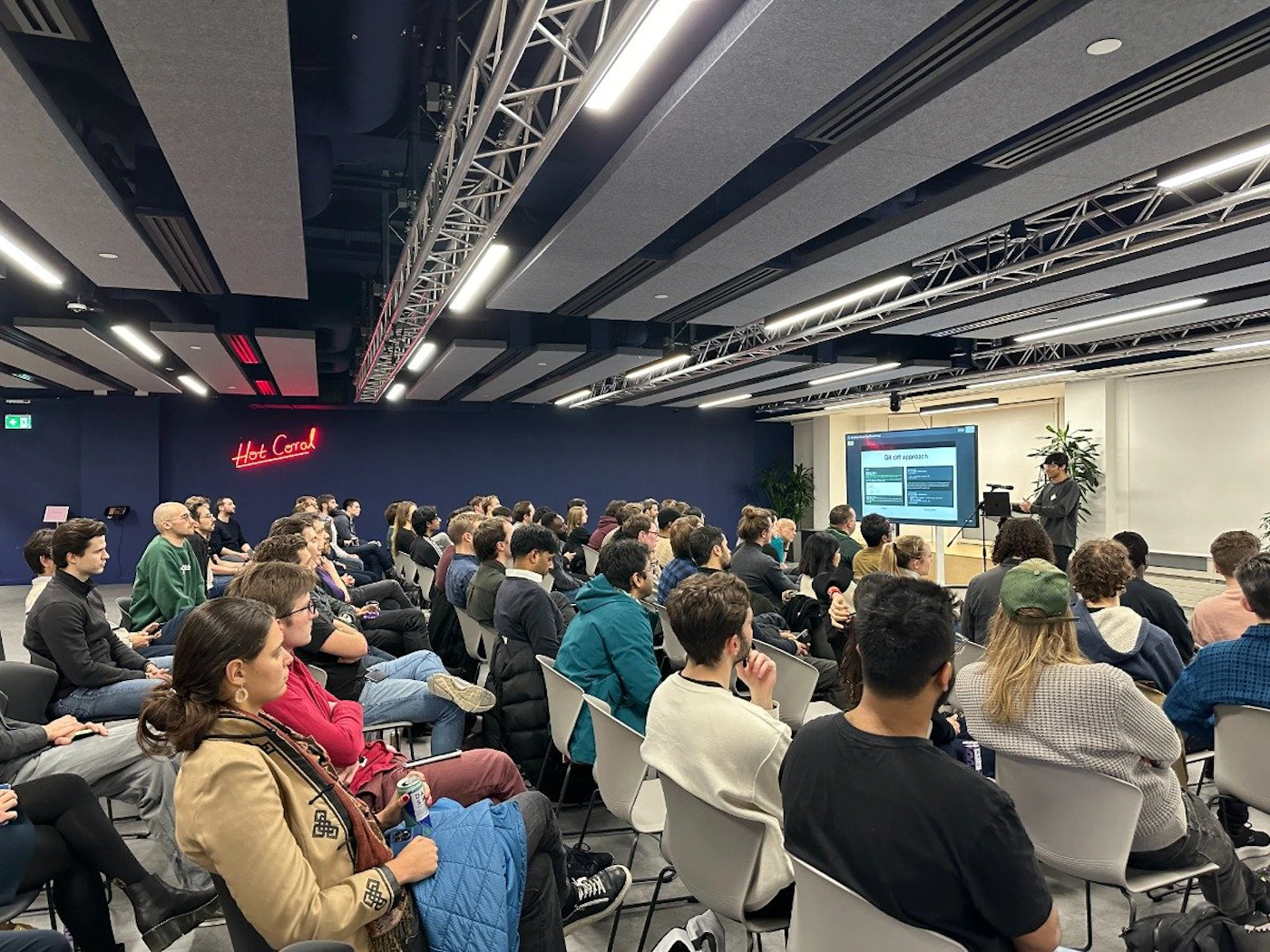 (Photo) A photo of a presentation taking place in a large room with an audience seated in chairs. Text: Hot Coral Git diff expression people | indoor | presentation, audience, screen | candid Note: The image is a real-life captured image of a presentation, depicting people, a venue, and a screen.  This clearly falls under the "photo" category.