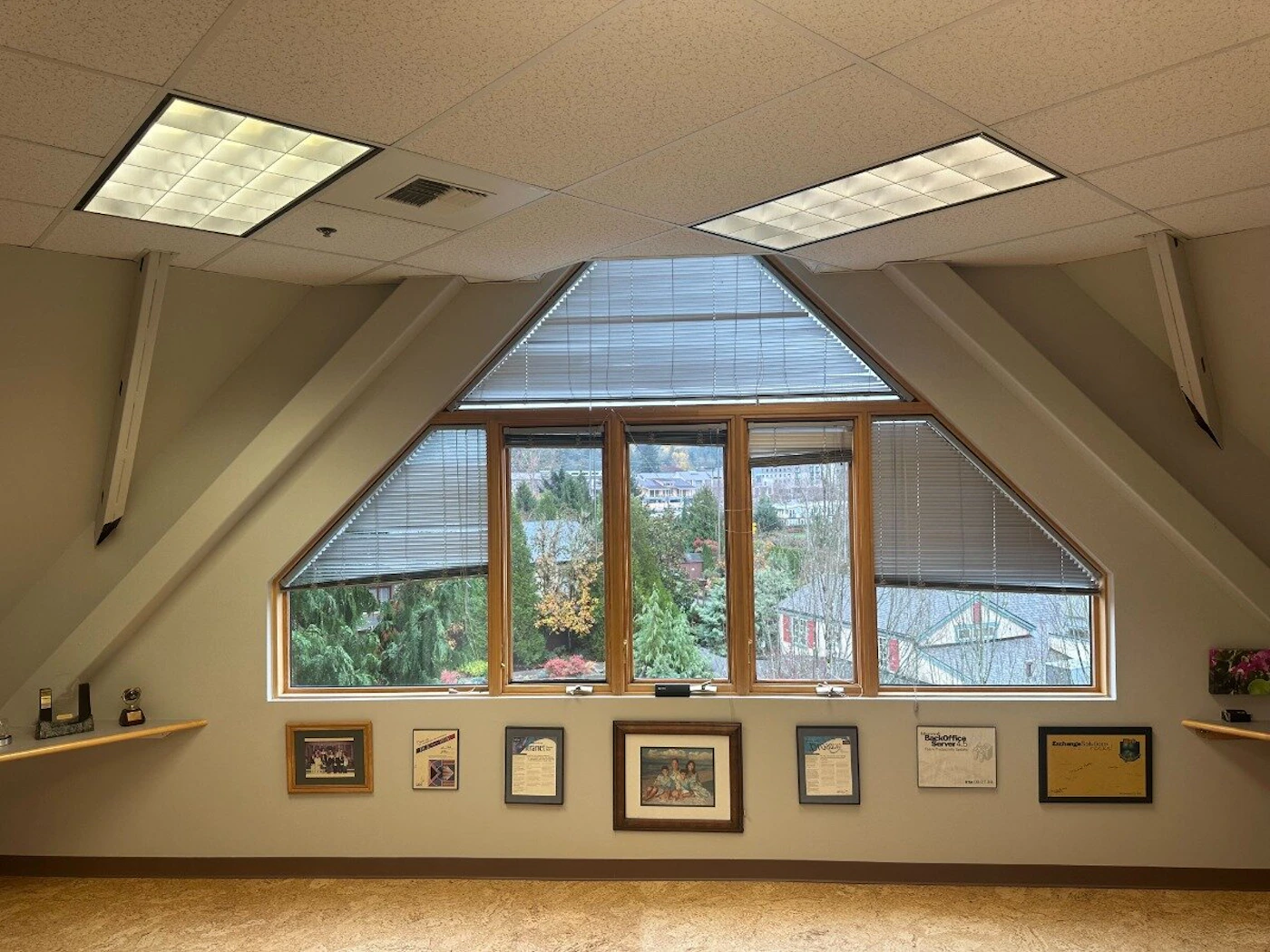 (Photo) An interior photograph of a room, likely an office space, featuring a distinctive triangular window structure that overlooks a residential area with dense greenery. Below the window, several framed documents and pictures are displayed on the beige wall. Interior architecture and office decor | Indoor room (attic/top floor office) with an outdoor view | Sloped ceiling, unique triangular window configuration, recessed fluorescent lighting, framed documents/awards on the wall. | Straight-on, architectural interior shot | Colors: #d9d2c1, #f5f5f5, #967355, #4c6e3d Note: The image is a realistic capture of a physical interior space, including architectural features, furniture, and a view of the outdoors, characteristic of a photograph.