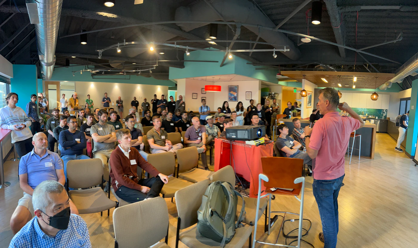 (Photo) A photo of a large group of people in a presentation or meeting setting, with one person appearing to speak to the audience. people | indoor | presentation, audience, projector | candid Note: The image is a real-world captured image of an event, depicting people and a setting. It has the characteristics of a photograph.